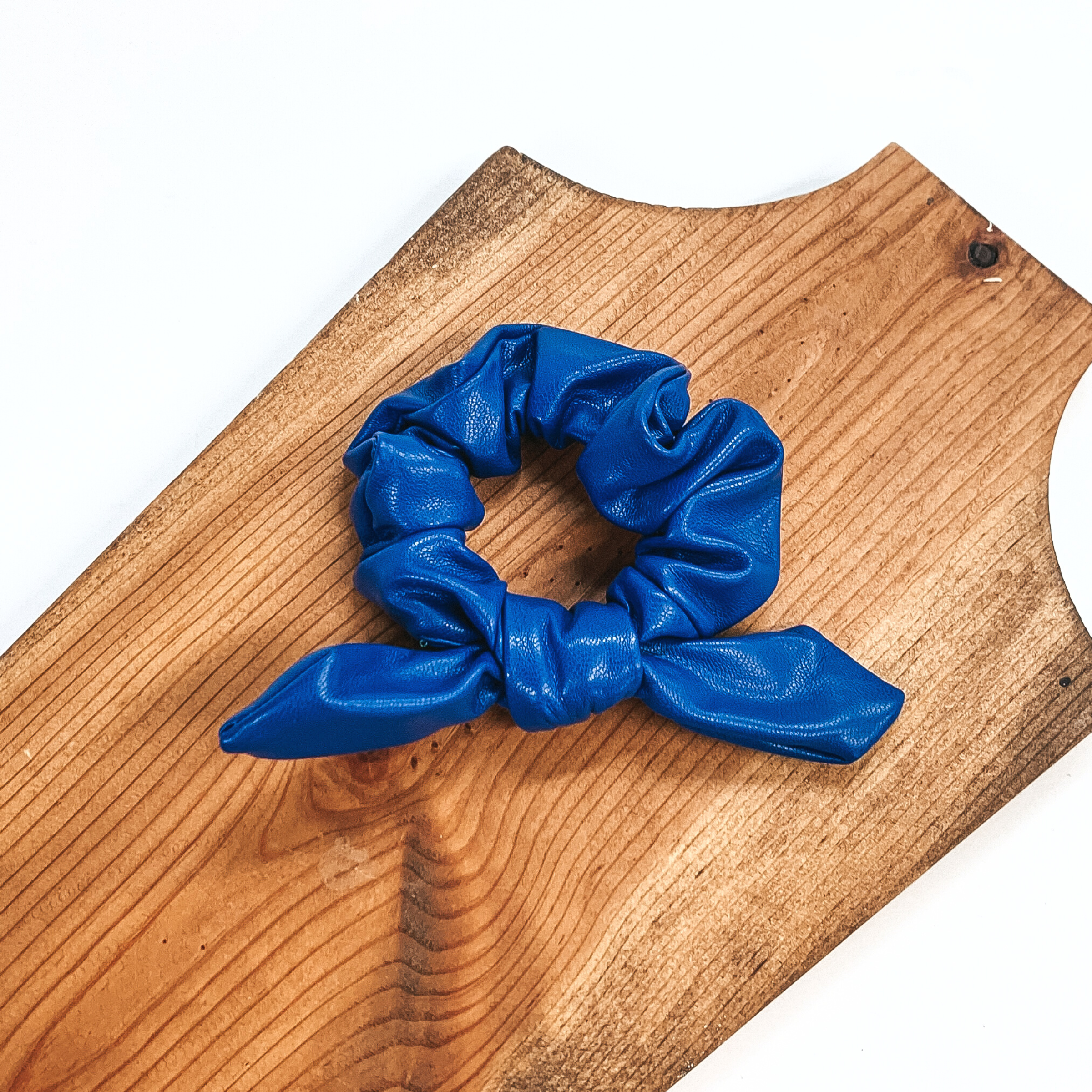 This is blue faux leather scrunchie with a bow, this scrunchie is taken on  a brown necklace board and on a white background.
