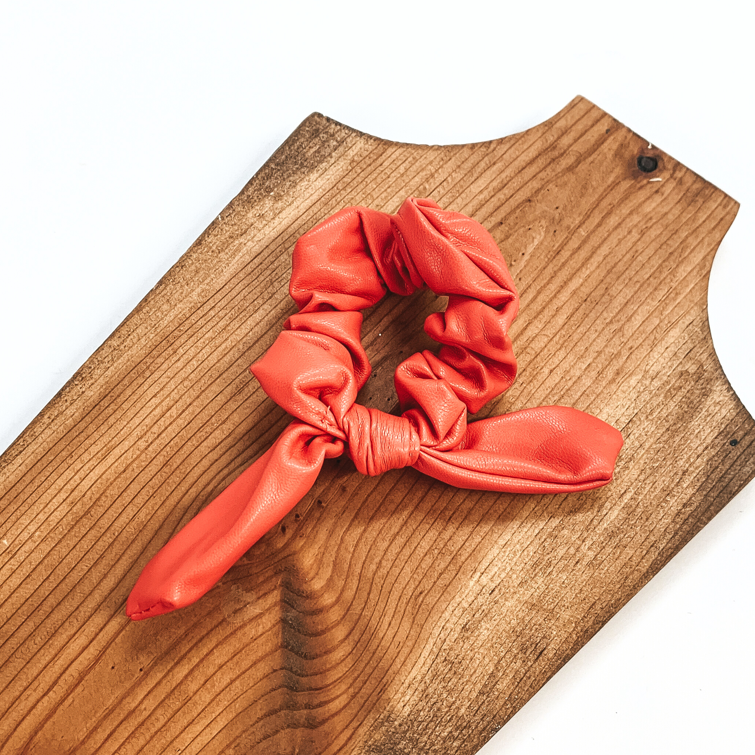 This is coral faux leather scrunchie with a bow, this scrunchie is taken on  a brown necklace board and on a white background.