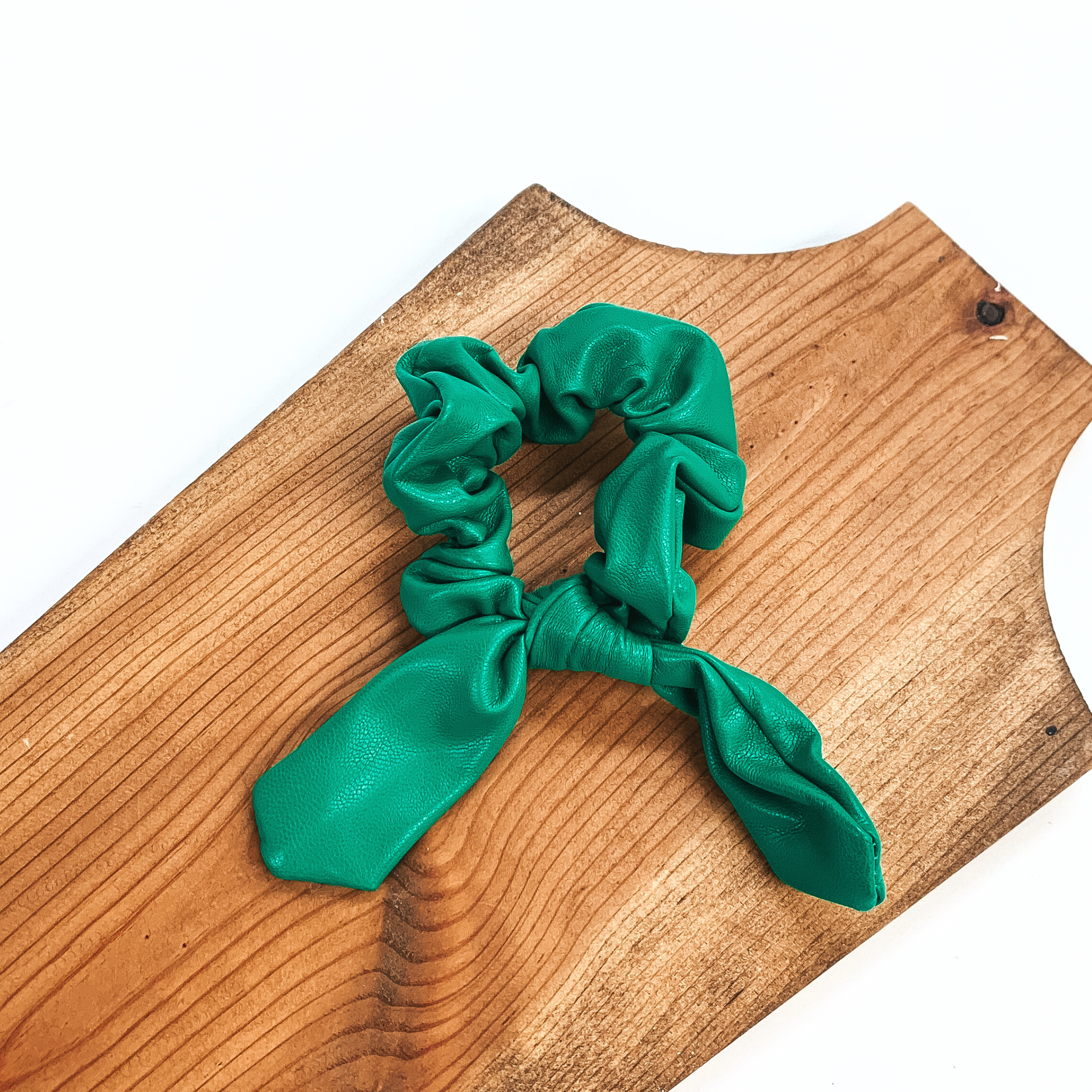 This is green faux leather scrunchie with a bow, this scrunchie is taken on  a brown necklace board and on a white background.