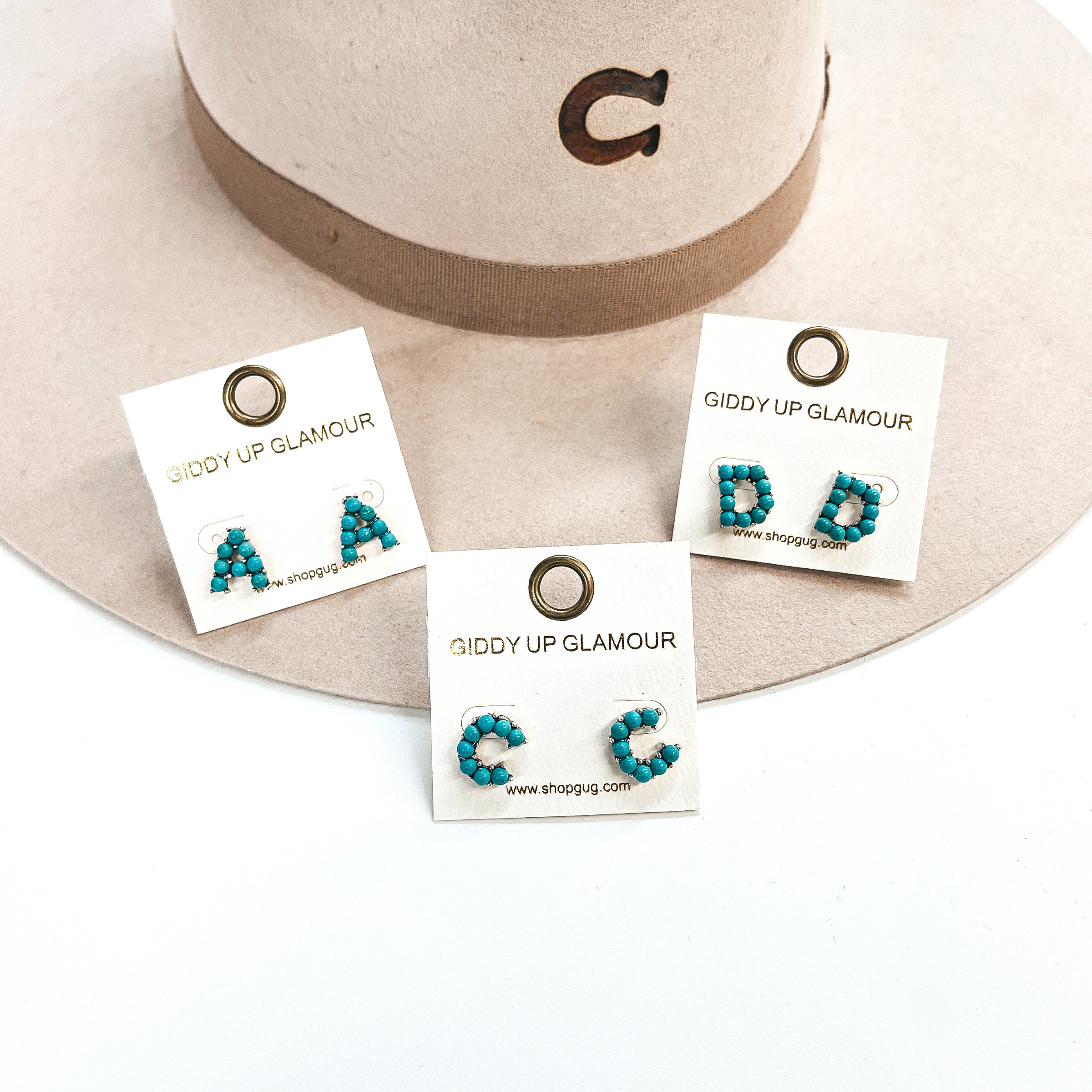 These are three pair of initial earrings with small turquoise stones in  a silver setting. From left to right; A,C,D. These earrings are placed  on a white Giddy Up Glamour card, they are laying on a beige, felt,  Charlie 1 Horse hat and on a white background.