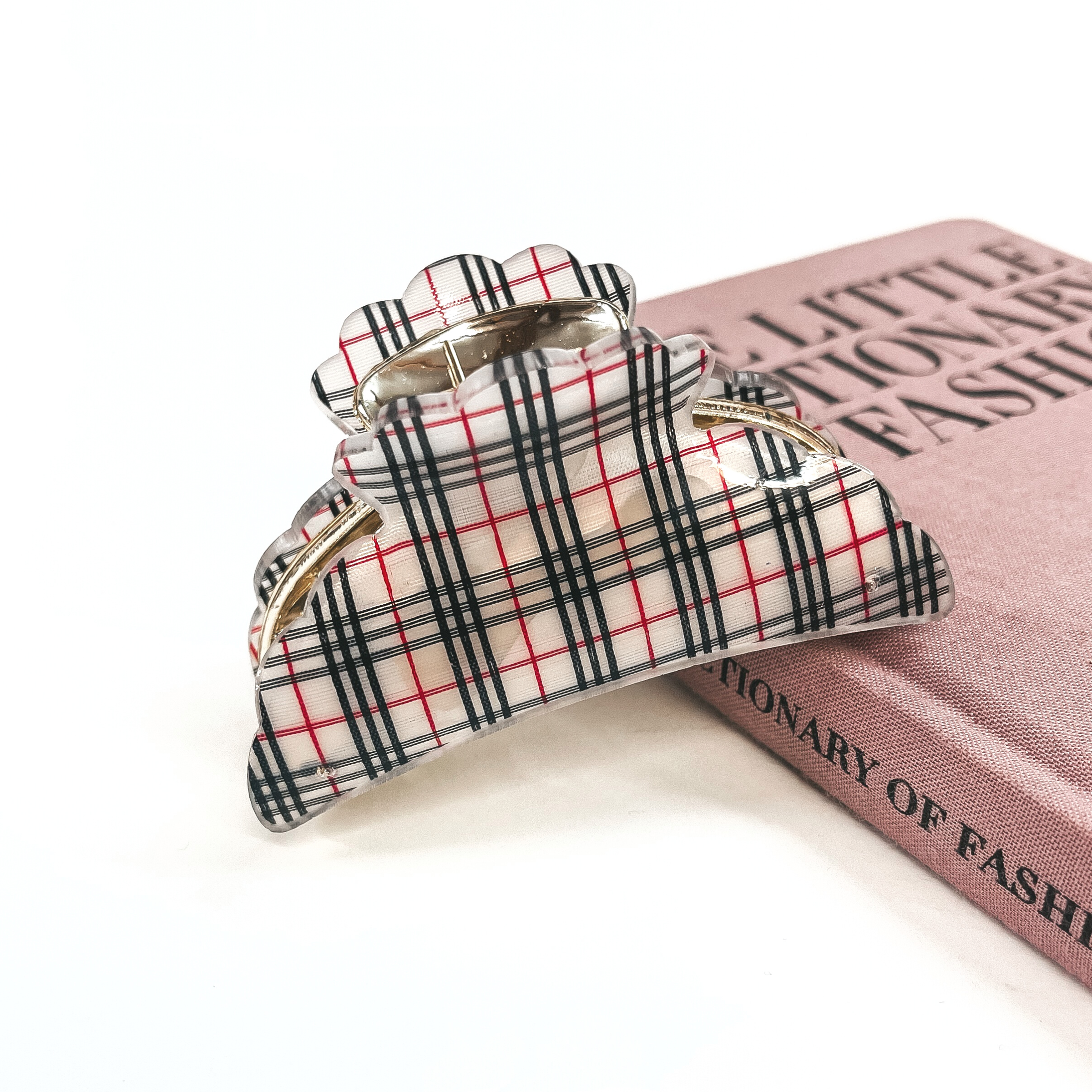 This is a gold and white plaid print hair clip laying on the side of a pink book and white background.