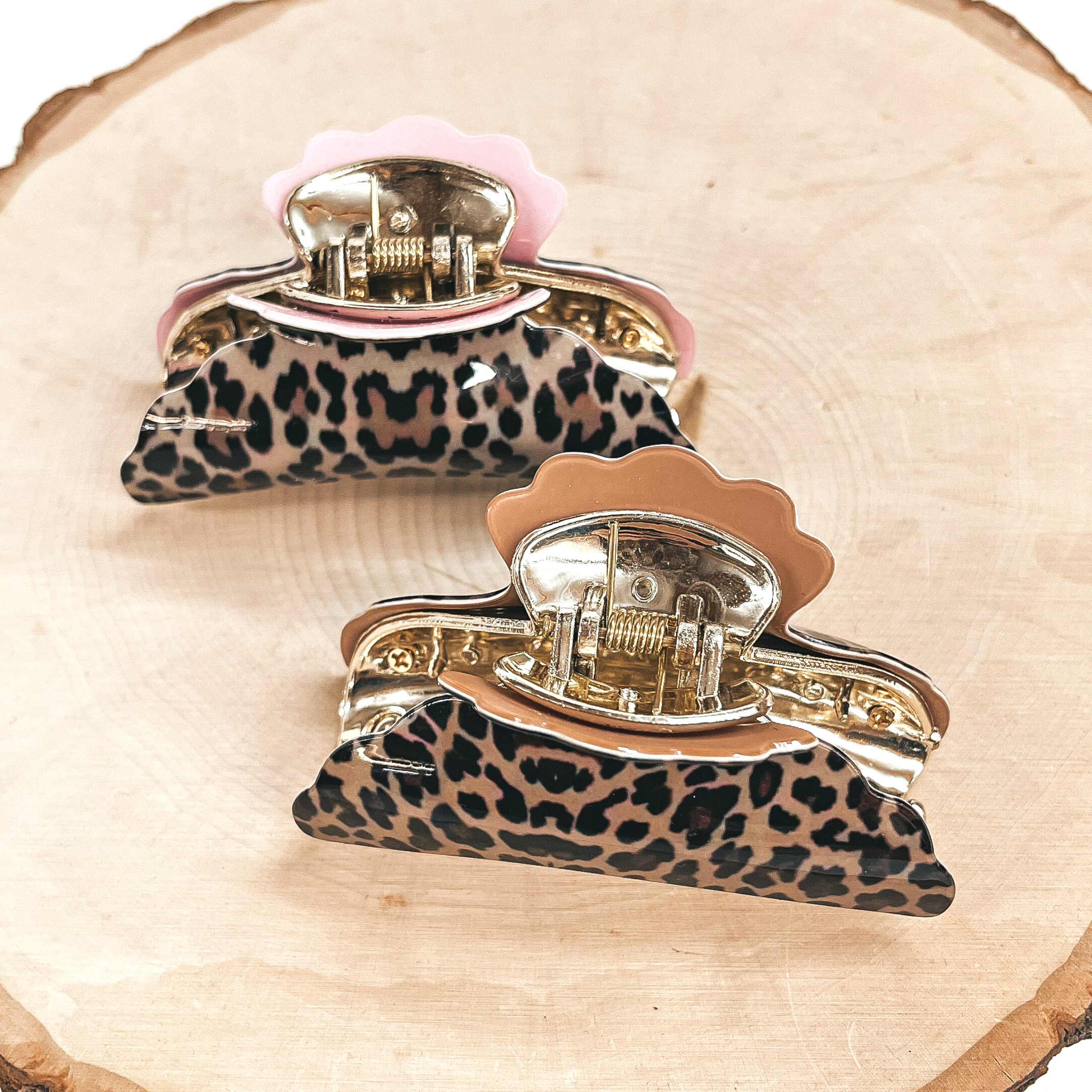 There are two light brown leopard print hair clips, the top one is in light pink and  the bottom one is in light brown, both are in a gold tone inlay. These hair clips  are taken on a slab of wood and white background.