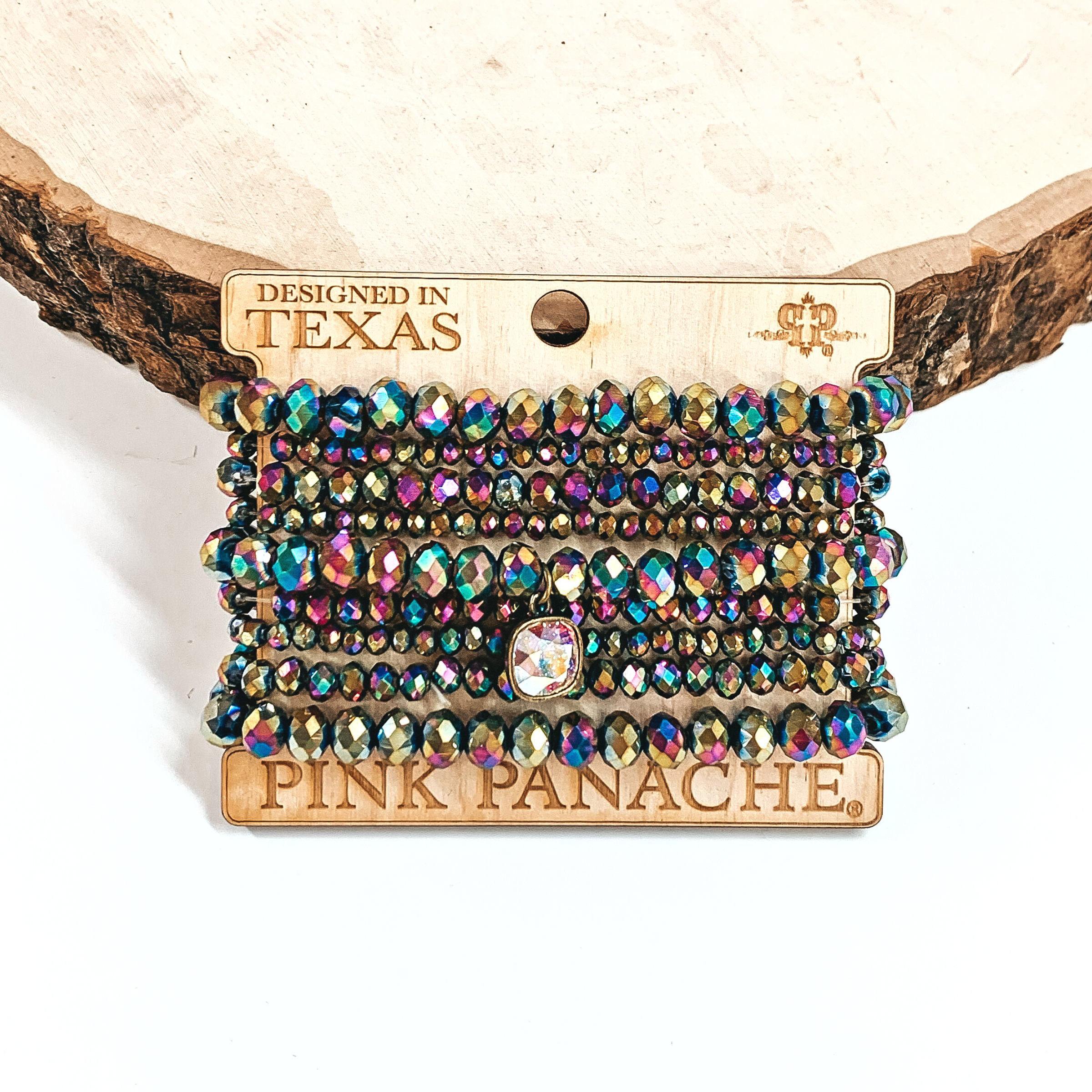 This is a nine set beaded bracelet in metallic rainbow with an AB cushion cut crystal  charm. The beaded bracelets have different sizes.  These bracelets are placed on a Pink Panache wooden holder, they are leaning up  against a slab of wood and on a white background.