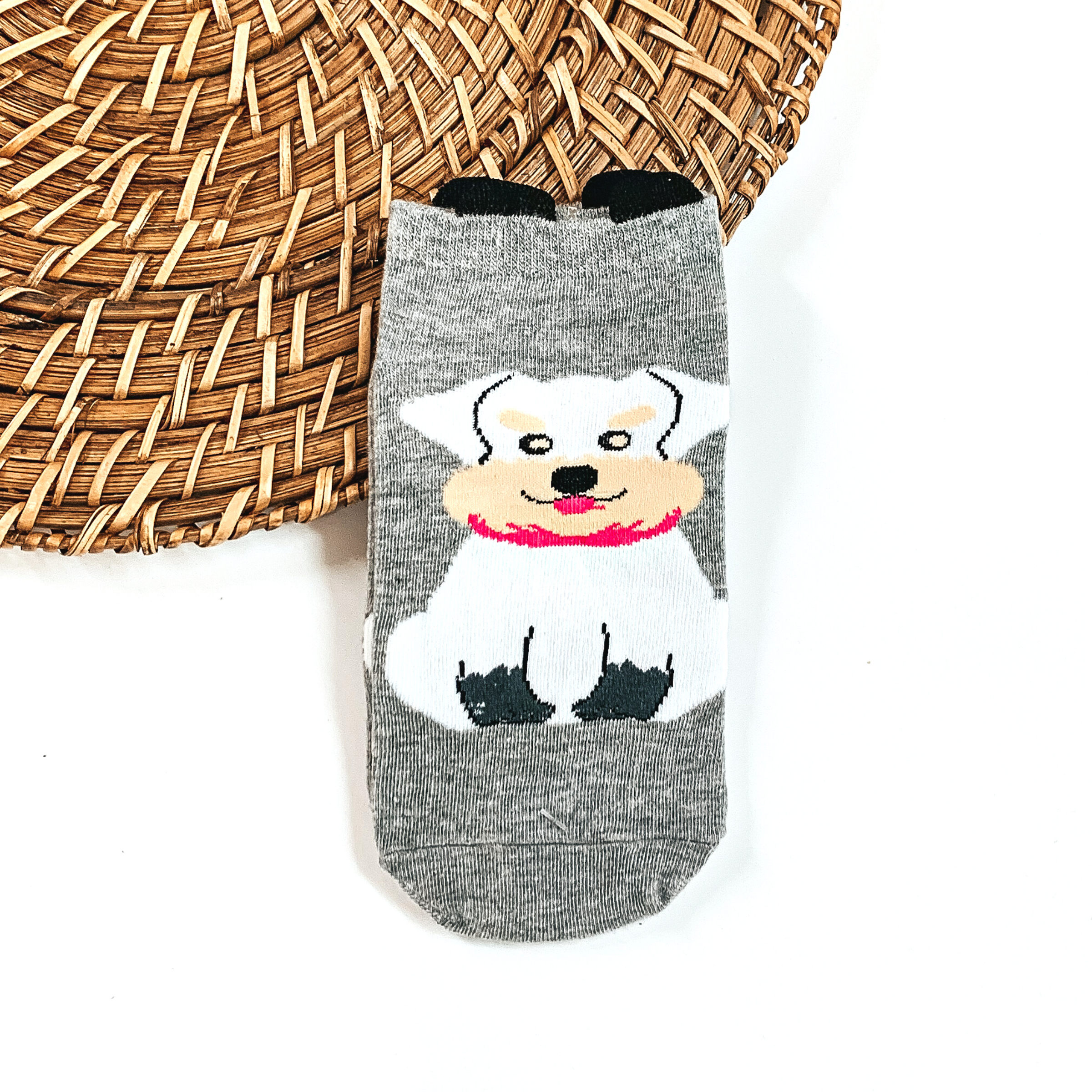 This is a grey pair of socks with a white Schnauzer dog and a pink scarf.  They have small black 'ears' on the top. This pair of socks are taken on a brown  woven plate and on a white background.