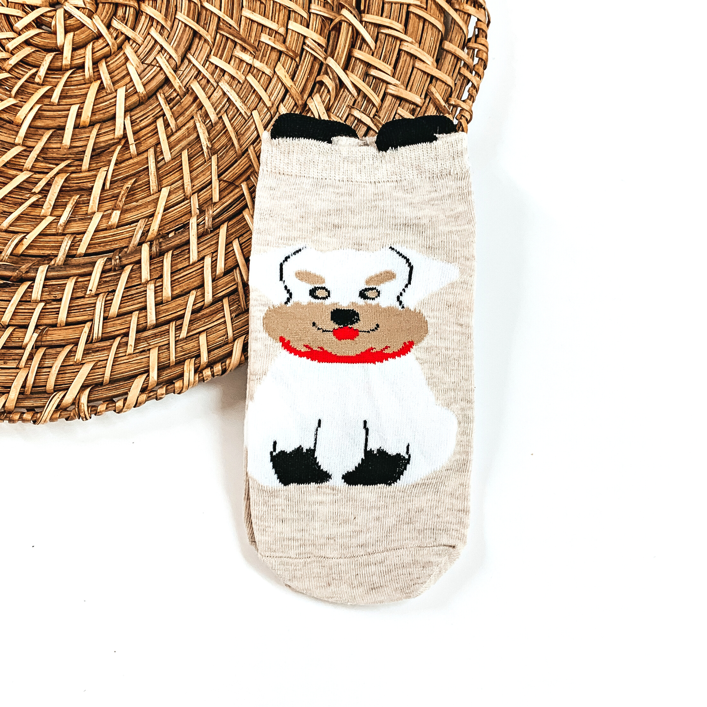 This is a beige pair of socks with a white Schnauzer dog and a red scarf.  They have small black 'ears' on the top. This pair of socks are taken on a brown  woven plate and on a white background.