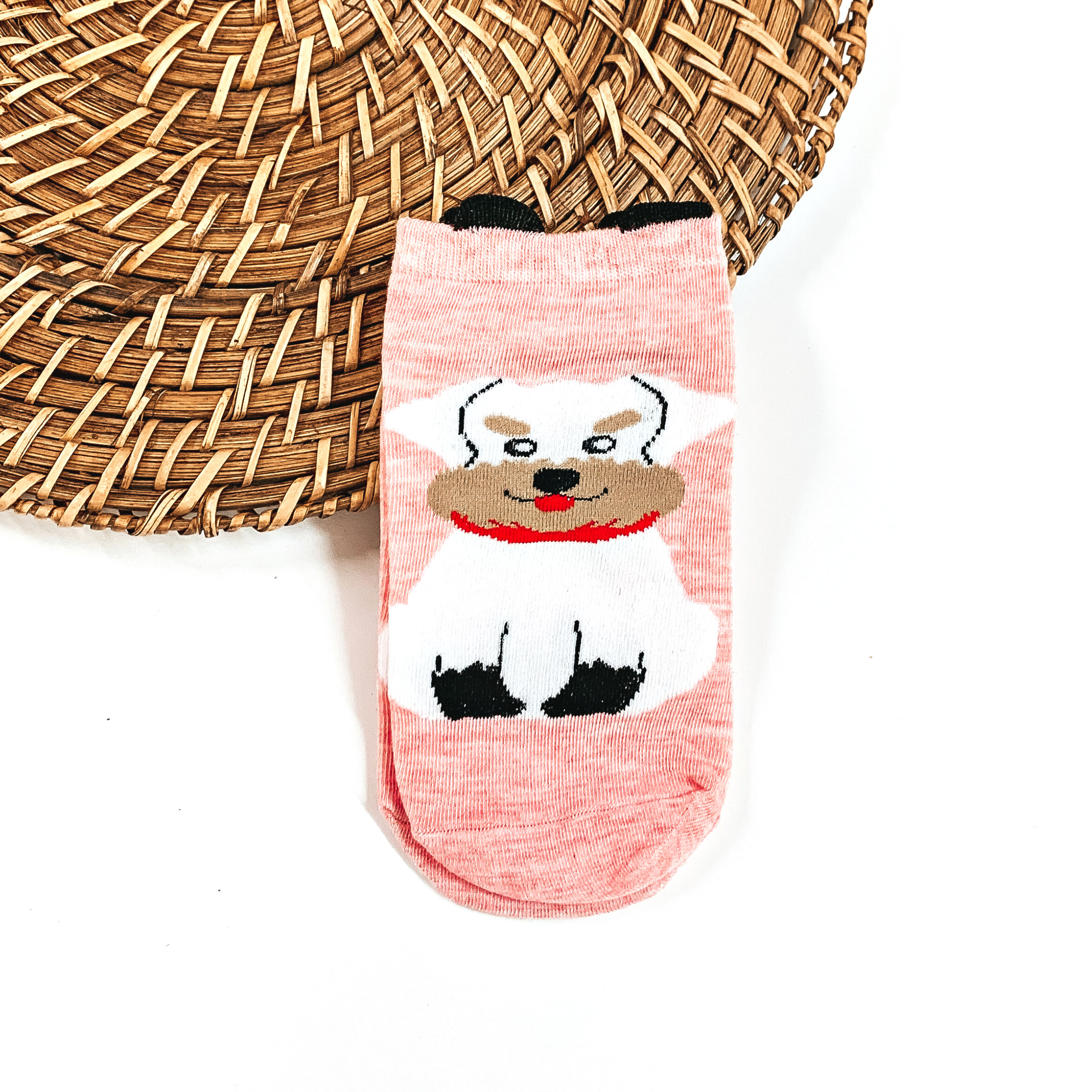 This is a light pink pair of socks with a white Schnauzer dog and a red scarf.  They have small black 'ears' on the top. This pair of socks are taken on a brown  woven plate and on a white background.