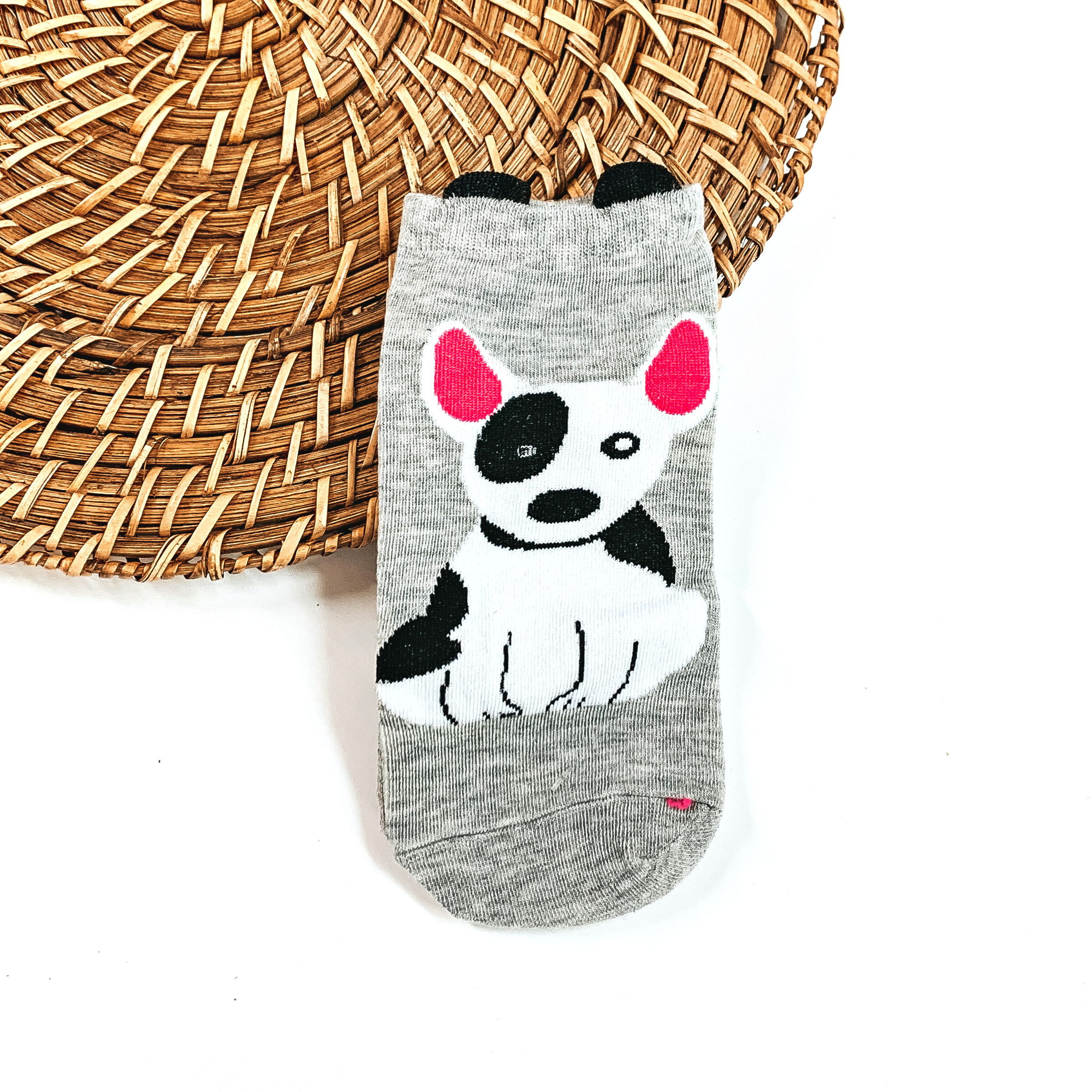 This is a grey pair of socks with a black/white Bull Terrier dog and pink ears. On top of the sock there are small black 'ears'. This pair of socks are taken on  a brown woven slate and on a white background.