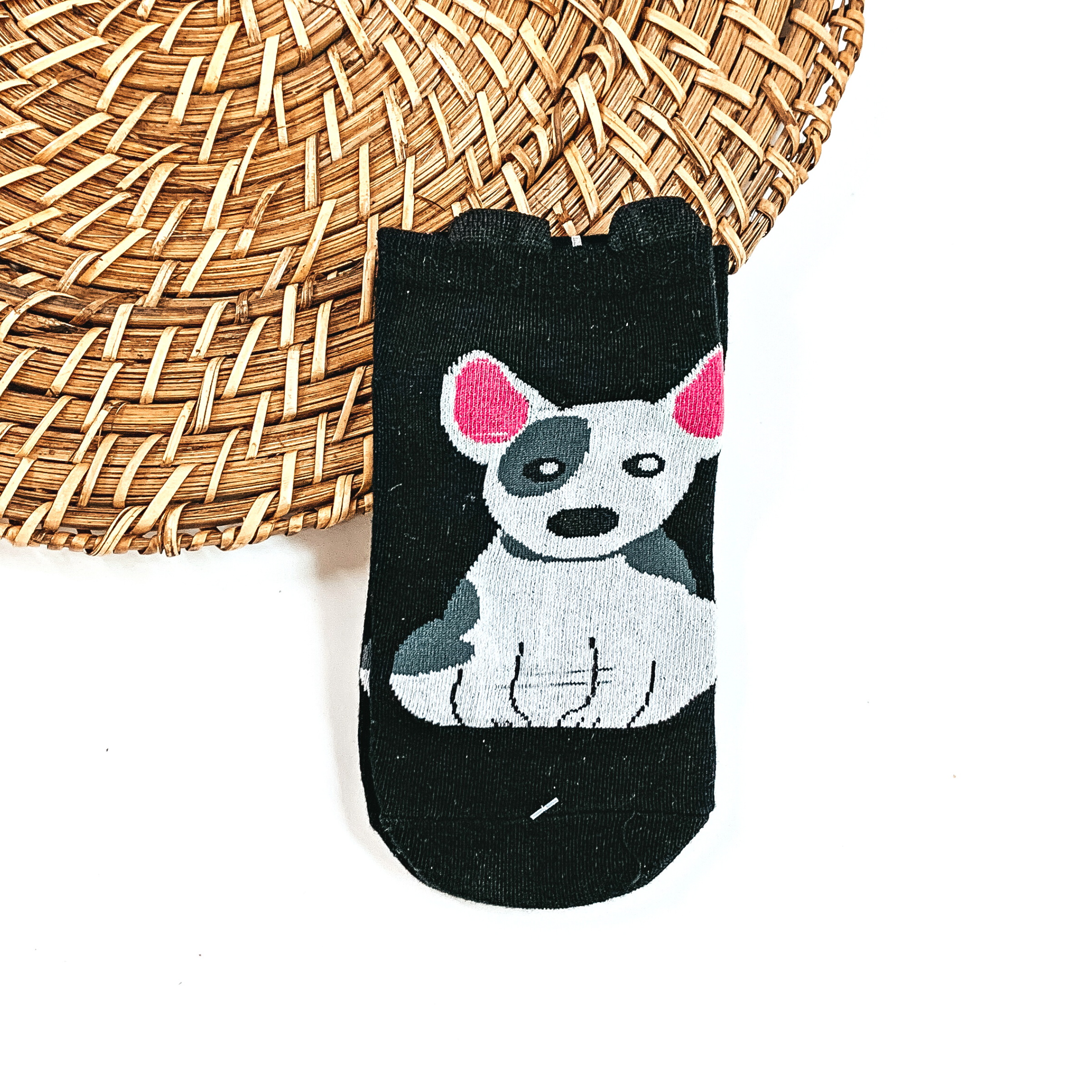 This is a black pair of socks with a grey/white Bull Terrier dog and pink ears. On top of the sock there are small black 'ears'. This pair of socks are taken on  a brown woven slate and on a white background.