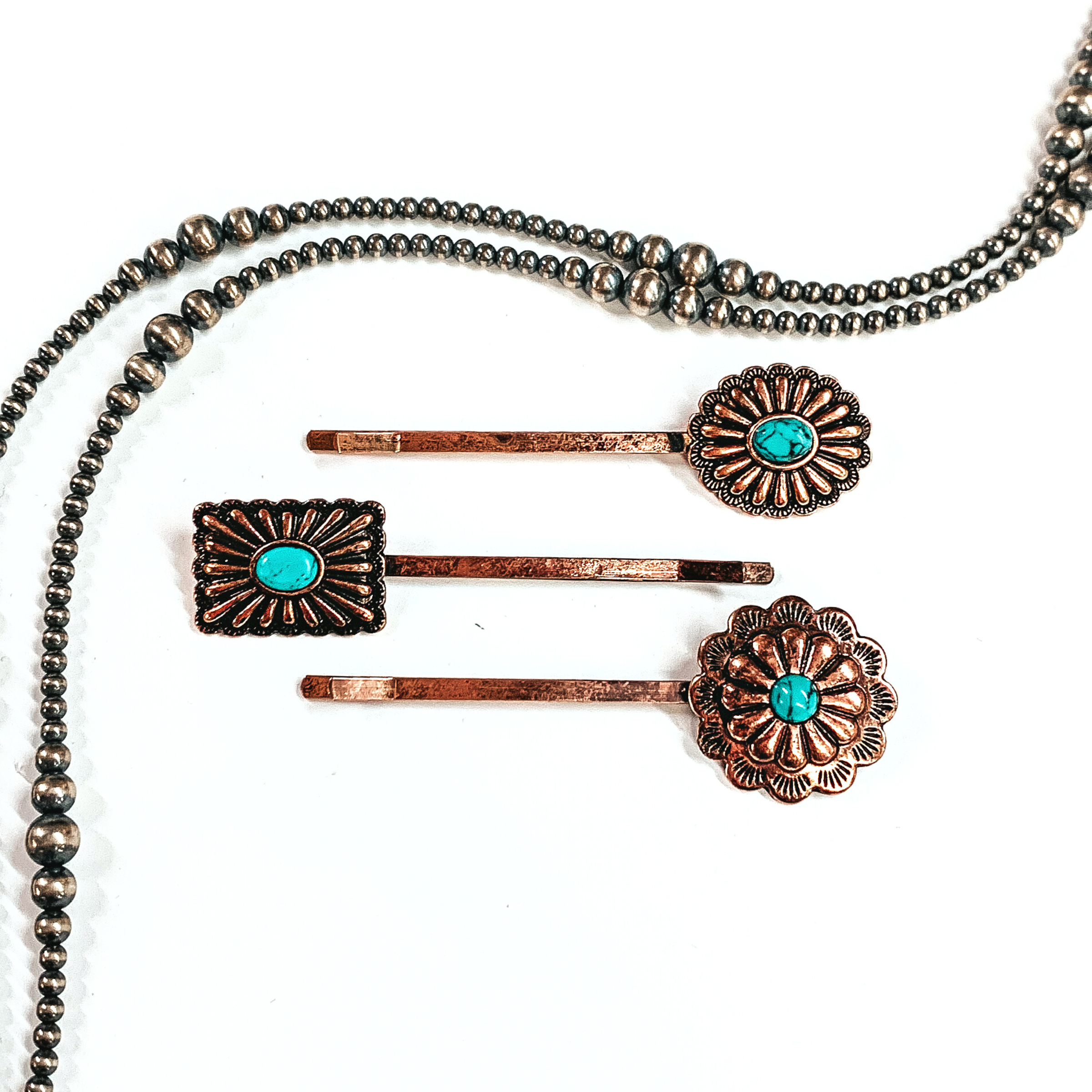 There are three copper bobby pins with conchos and a small faux turquoise stones  in the center of the concho. From top to bottom; an oval concho, rectangle/square  concho, and then a flower concho. These bobby pins are taken on a white background  with silver navajo pearls around as decor.