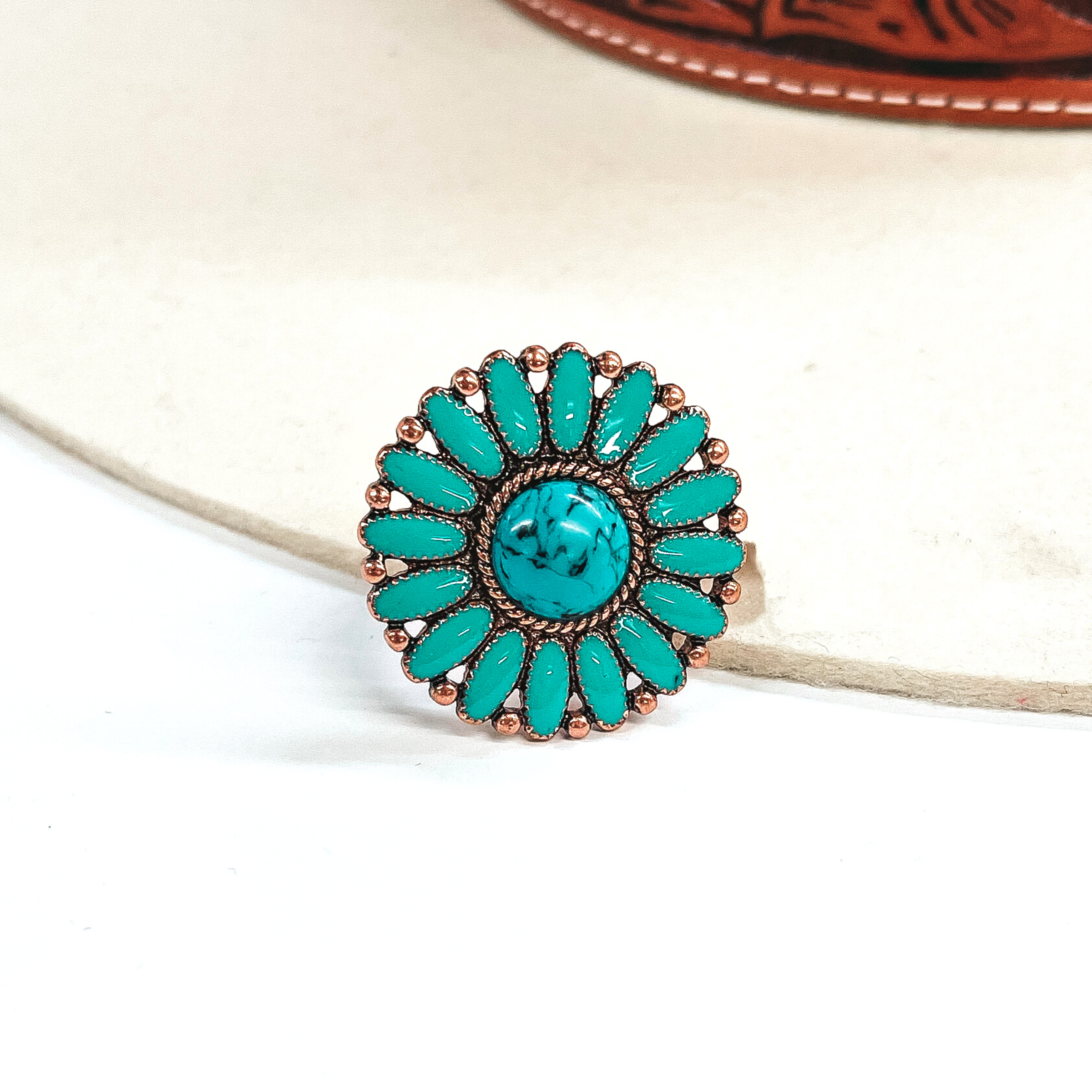 This is a turquoise,circle concho, epoxy cluster ring in a copper setting.  The ring has a small circle stone in the center with a copper, rope textured outline,  with oval shaped stone all around like a flower. In between each 'petal' it has copper  circles all around. This ring is taken on an ivory felt hat  with a brown textured band and on a white background.