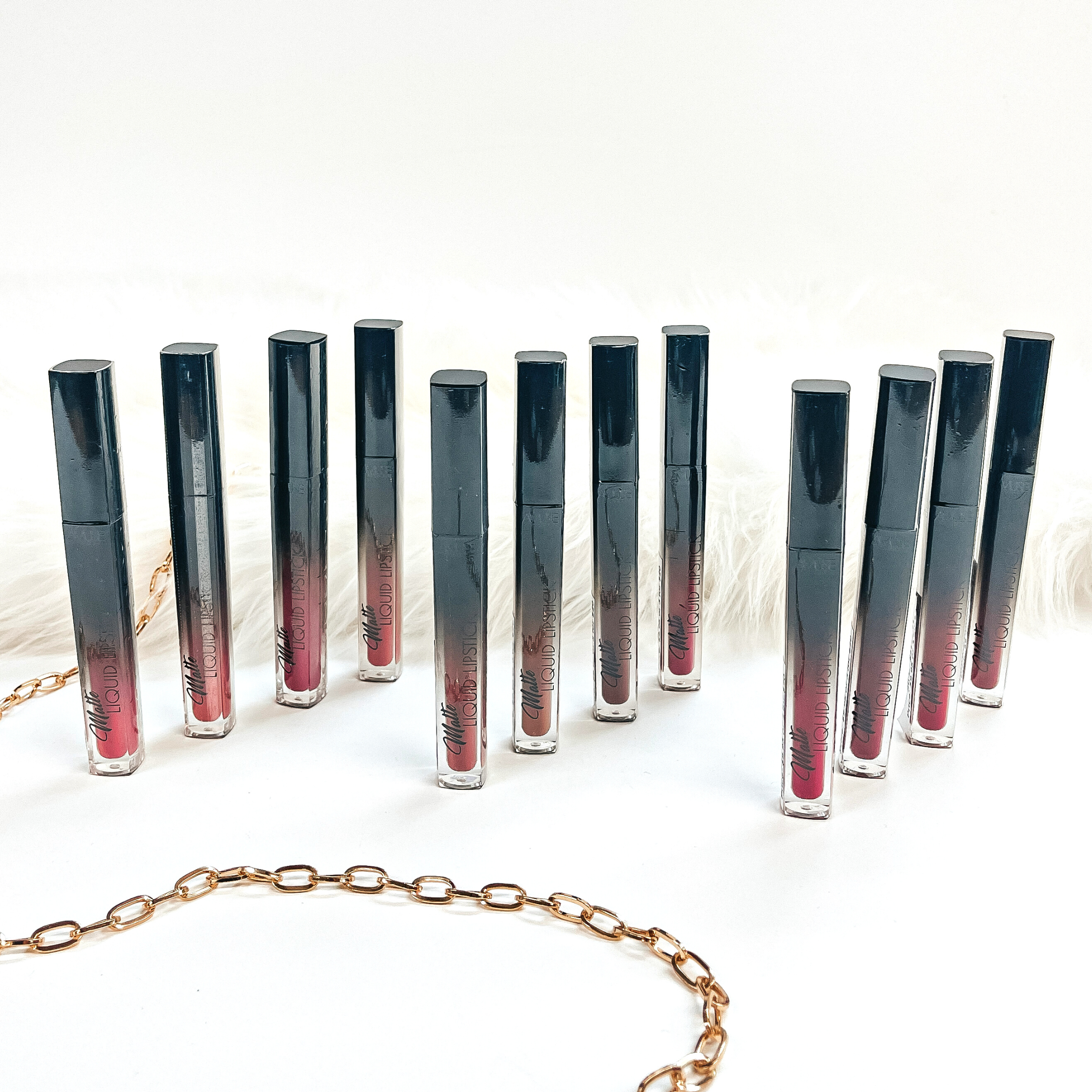 There are twelve liquid lipsticks in different shades of pink/nudes/reds.  These lipsticks are taken in groups of four, making three diagonal rows. They  are taken on a white background with white fur in the back and a gold link chain  as decor.