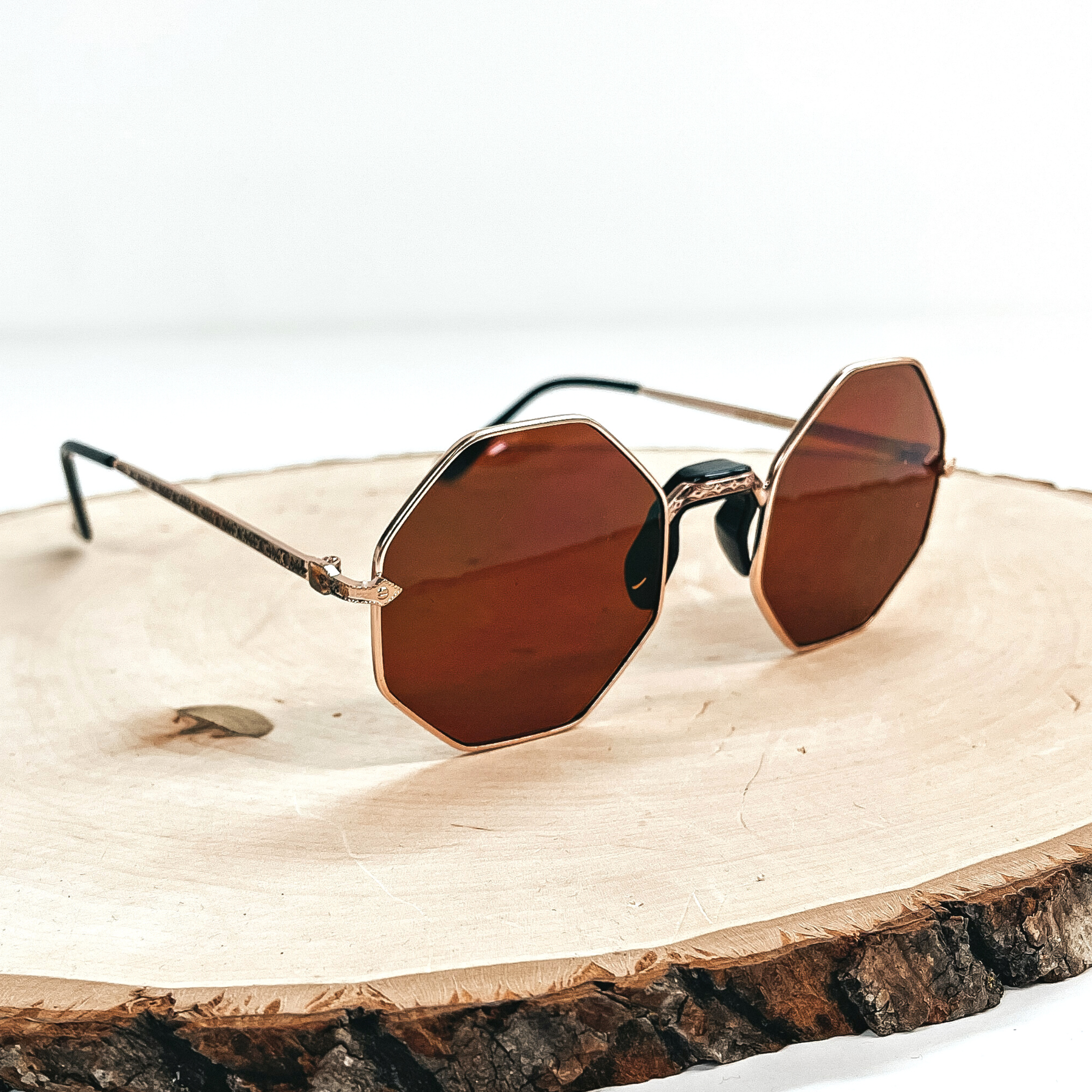 This is a pair of brown octagon sunglasses, the lense is brown with a  rose gold outline and a black nose piece. These pair of sunglasses are taken on a wooden slate and on a white  background.