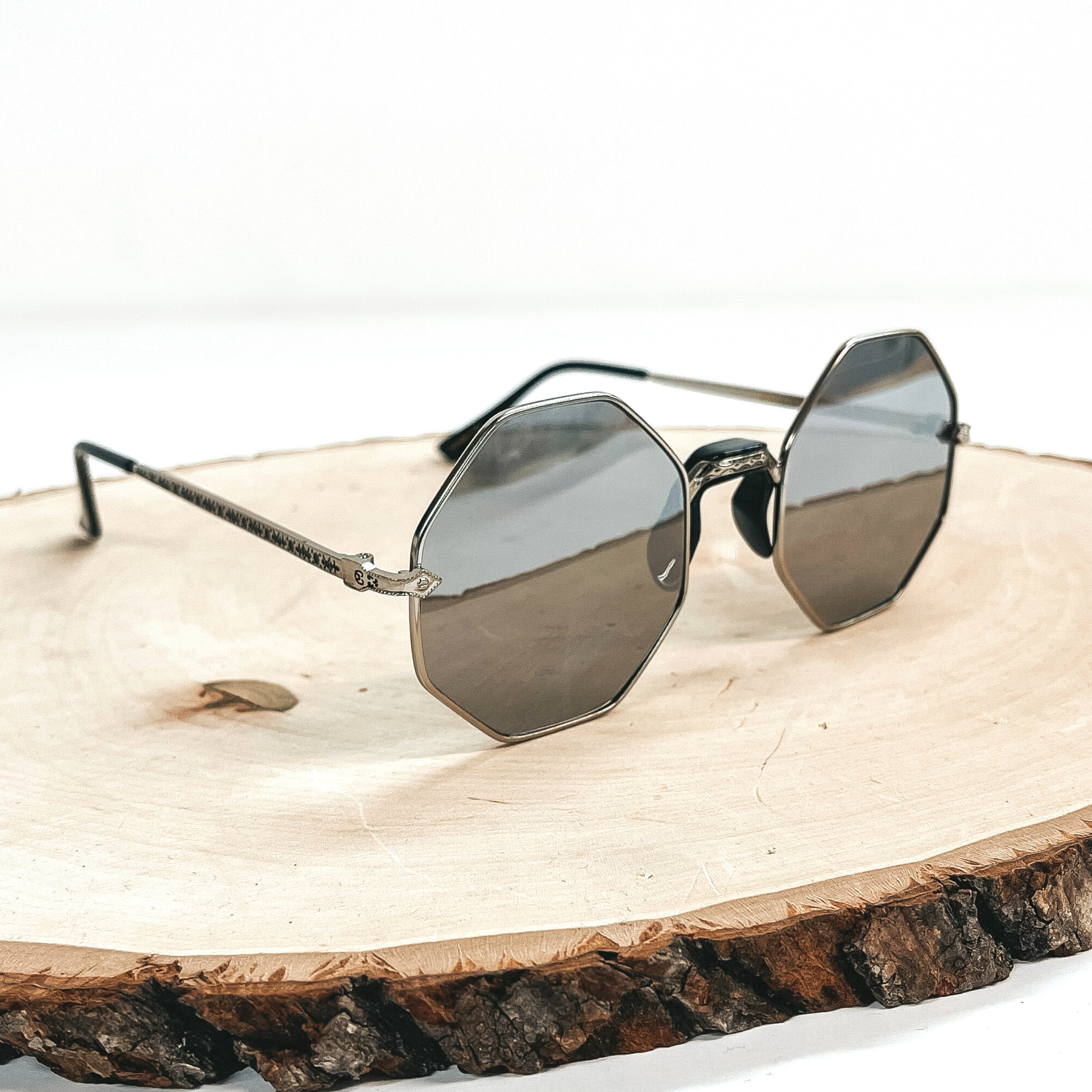 This is a pair of silver octagon sunglasses, the lense is silver/grey with a  silver outline and a black nose piece. These pair of sunglasses are taken on a wooden slate and on a white  background.
