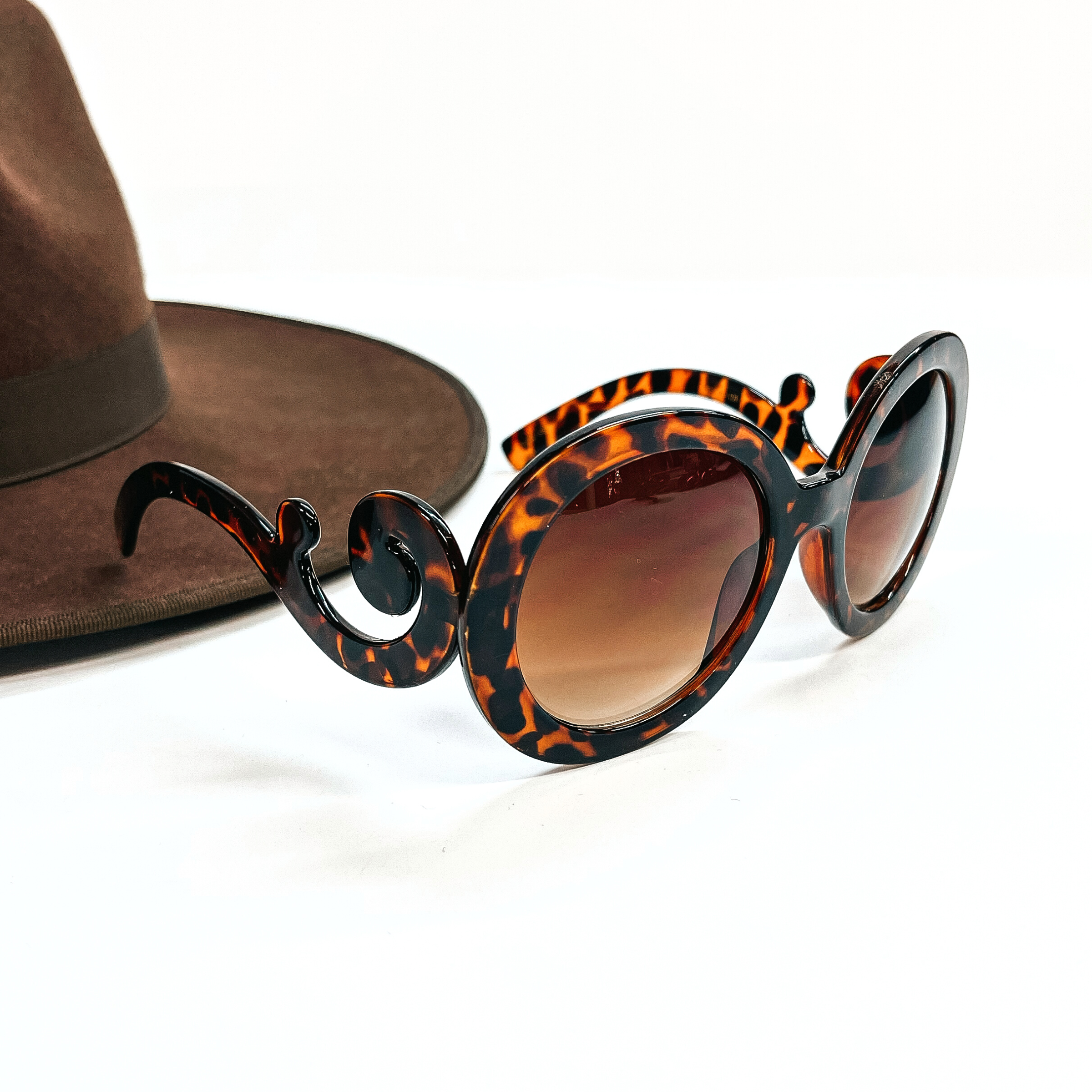 This is a brown pair of sunglasses, the frame is tortouise print with a  brown lense. The side parts of the sunglasses have a swirl. These sunglasses are taken on a white  background with a dark brown felt hat in the side as decor.