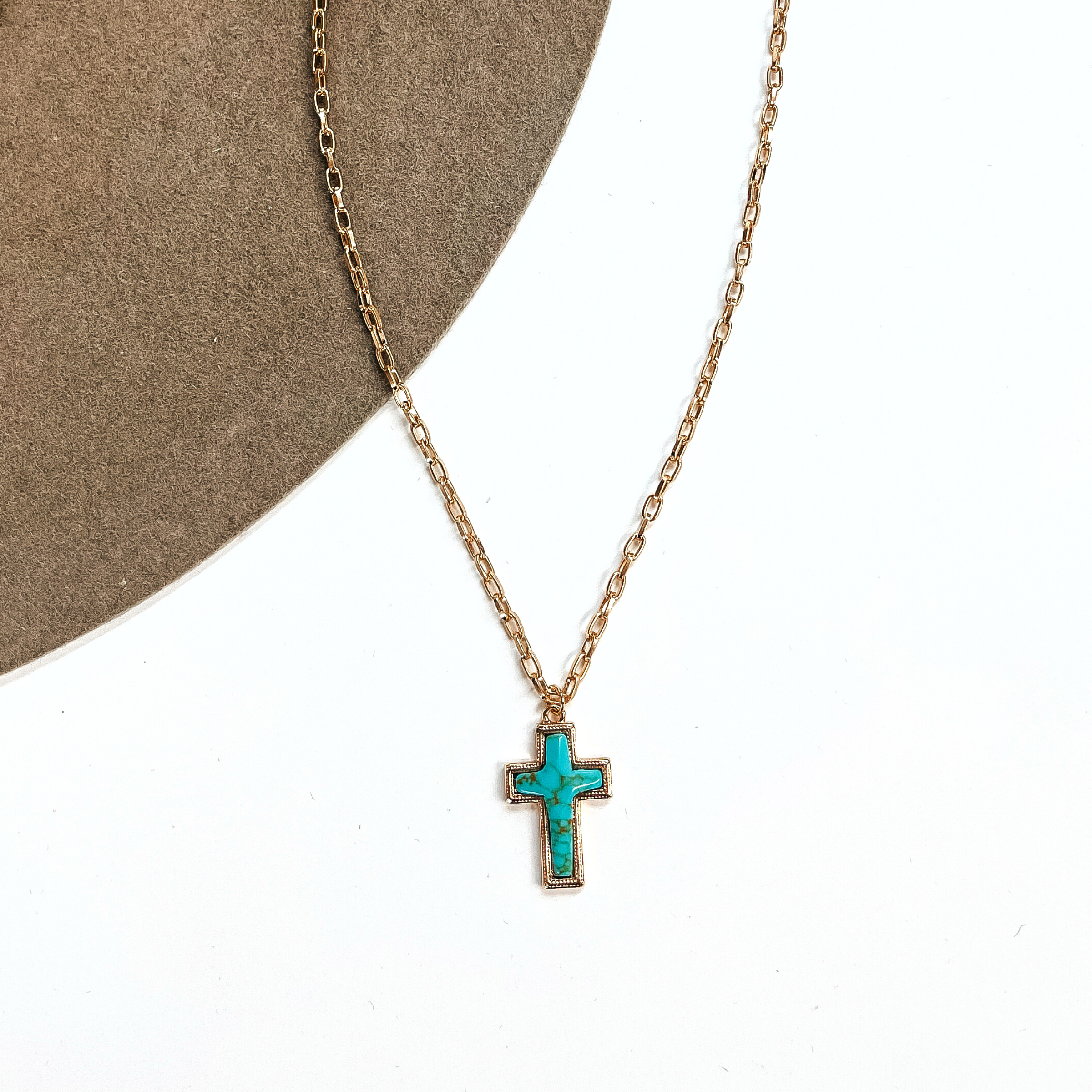 This is a thin gold tone chain necklace with a small cross pendant in the center. The  pendant has a turqouise stone shaped as a cross, the stone is in a gold setting. This  necklace is laying on a white background and on a light brown felt hat brim.