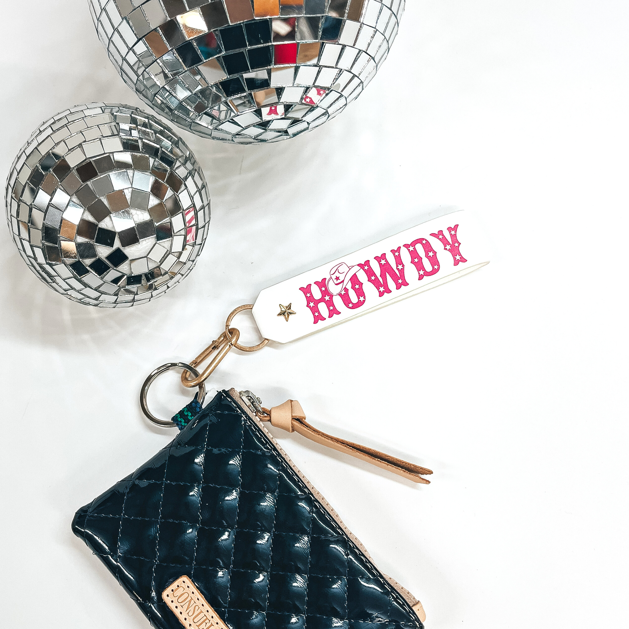 This is a white colored leather keychain with a matte gold key ring.  The keychain says, Howdy, in uppercase with star print inside and a  cowboy hat on top of the, O, in hot pink. It's connected to a black shiny Consuela pouch and on a white background with  two disco balls in the back as decor.