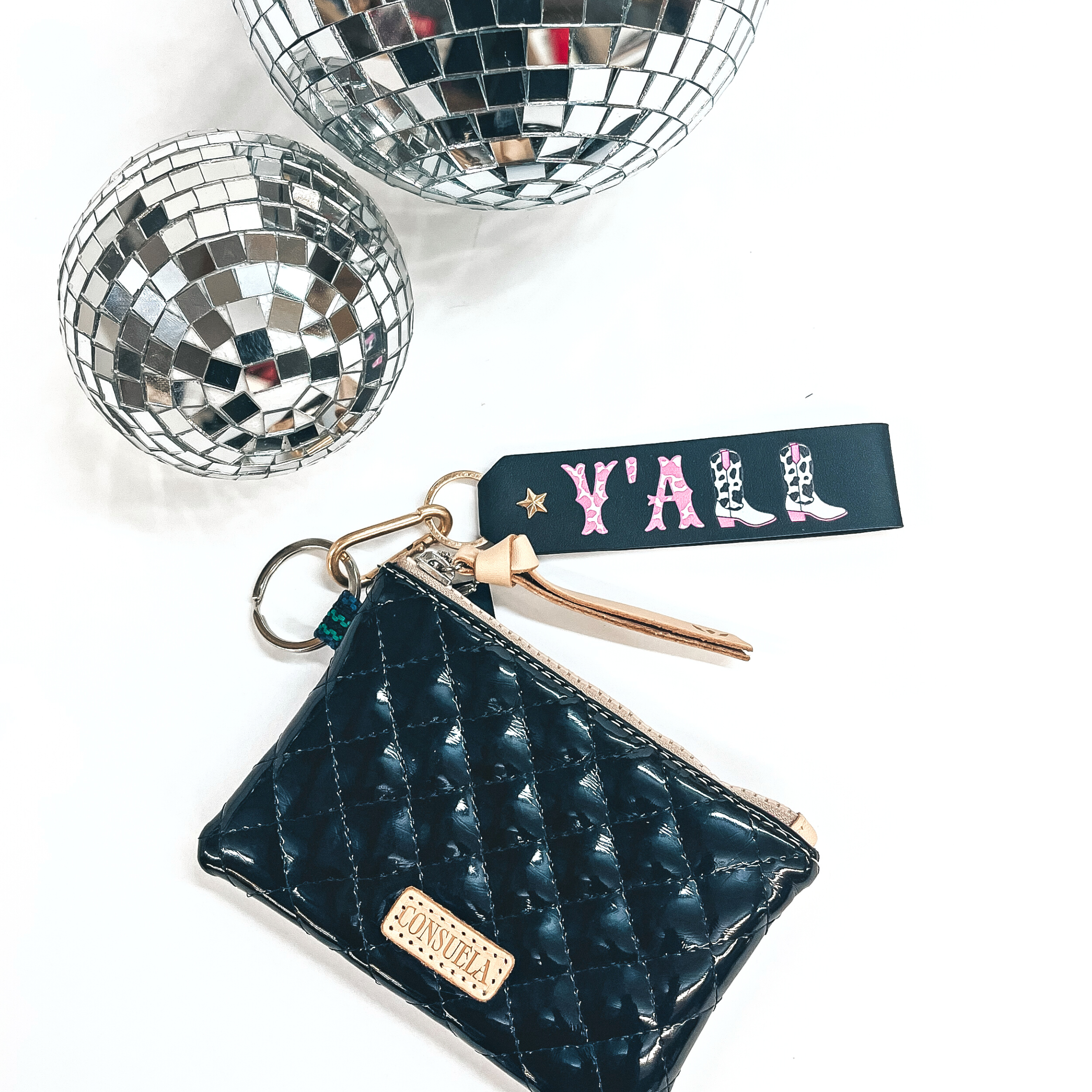 This is a black leather keychain with a matte gold key ring and a star.  It says, 'Y'ALL', with pink and white cow print inside the letters, there is  also two boots as L's. The boots have black and white cow print with pink  detailing. This keychain is connected to a black shiny Consuela pouch and on a  white background with two disco balls in the back as decor.