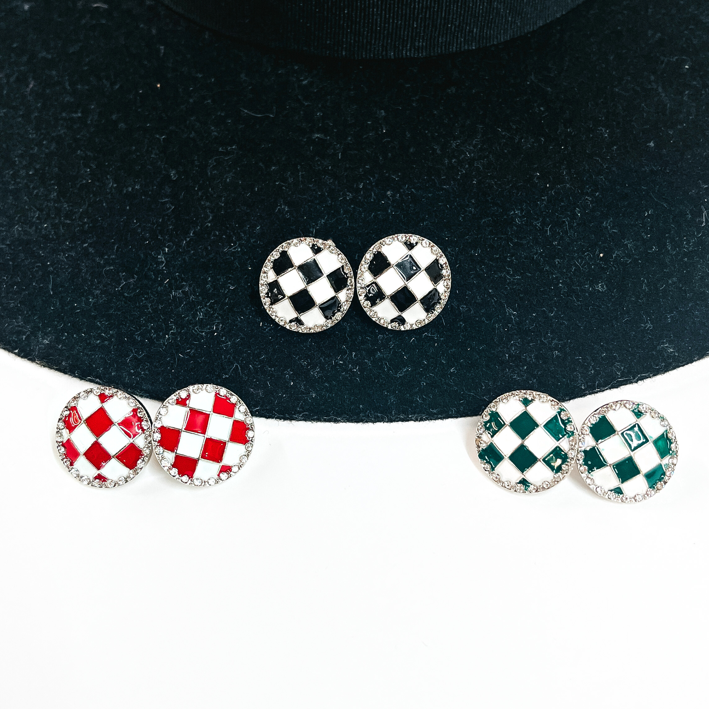 There three pairs of checkered pattern stud earrings in differet colors. From left to right; red/white, black/white, and green/white. These earrings are in a silver setting and have clear crystals all around. These pairs of earrings are laying on a black felt hat brim and on white background.