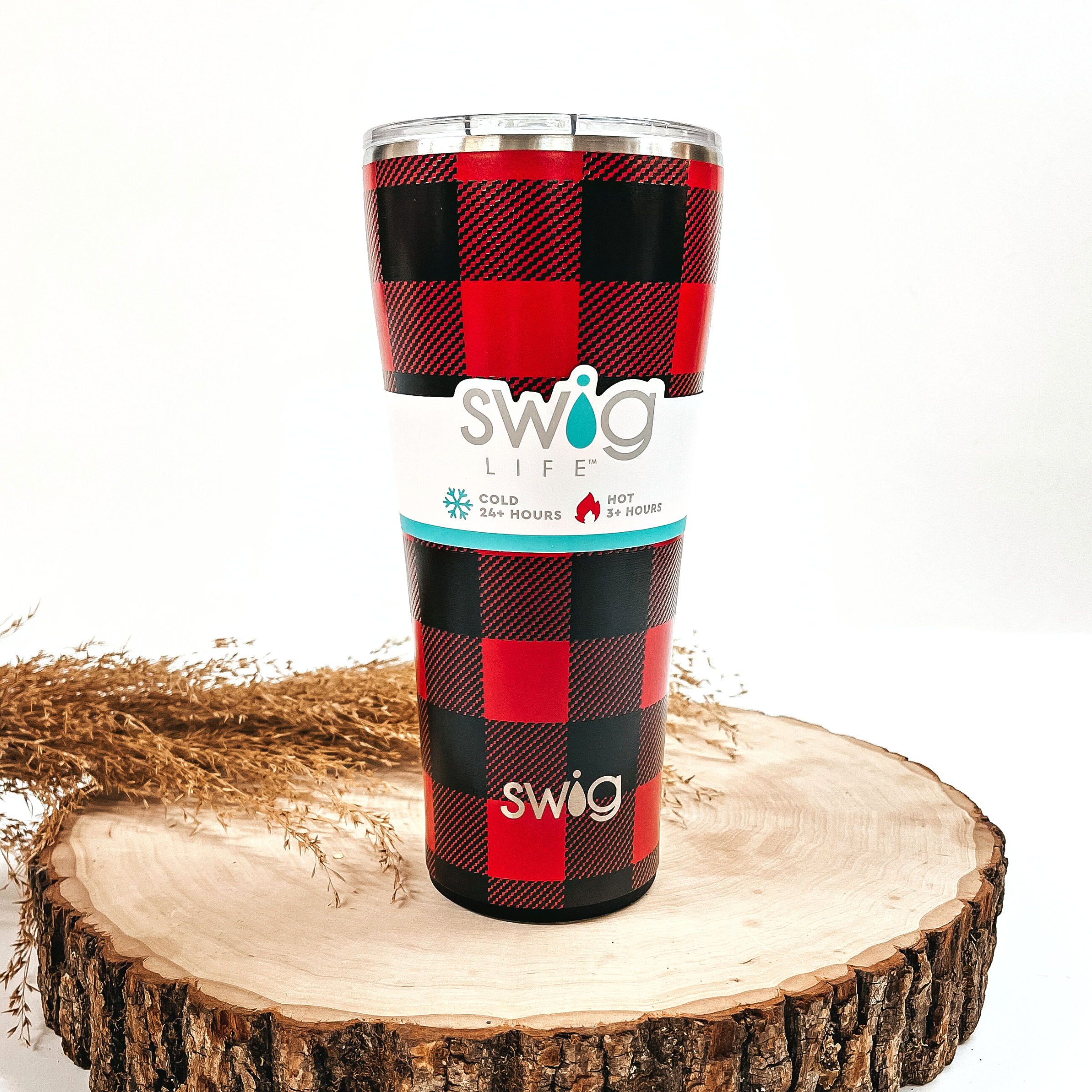 This is a red buffalo plaid cup in red and black, with a silver metallic top. This cup is taken on top of a slab of wood with a brown plant in the side  as decor.
