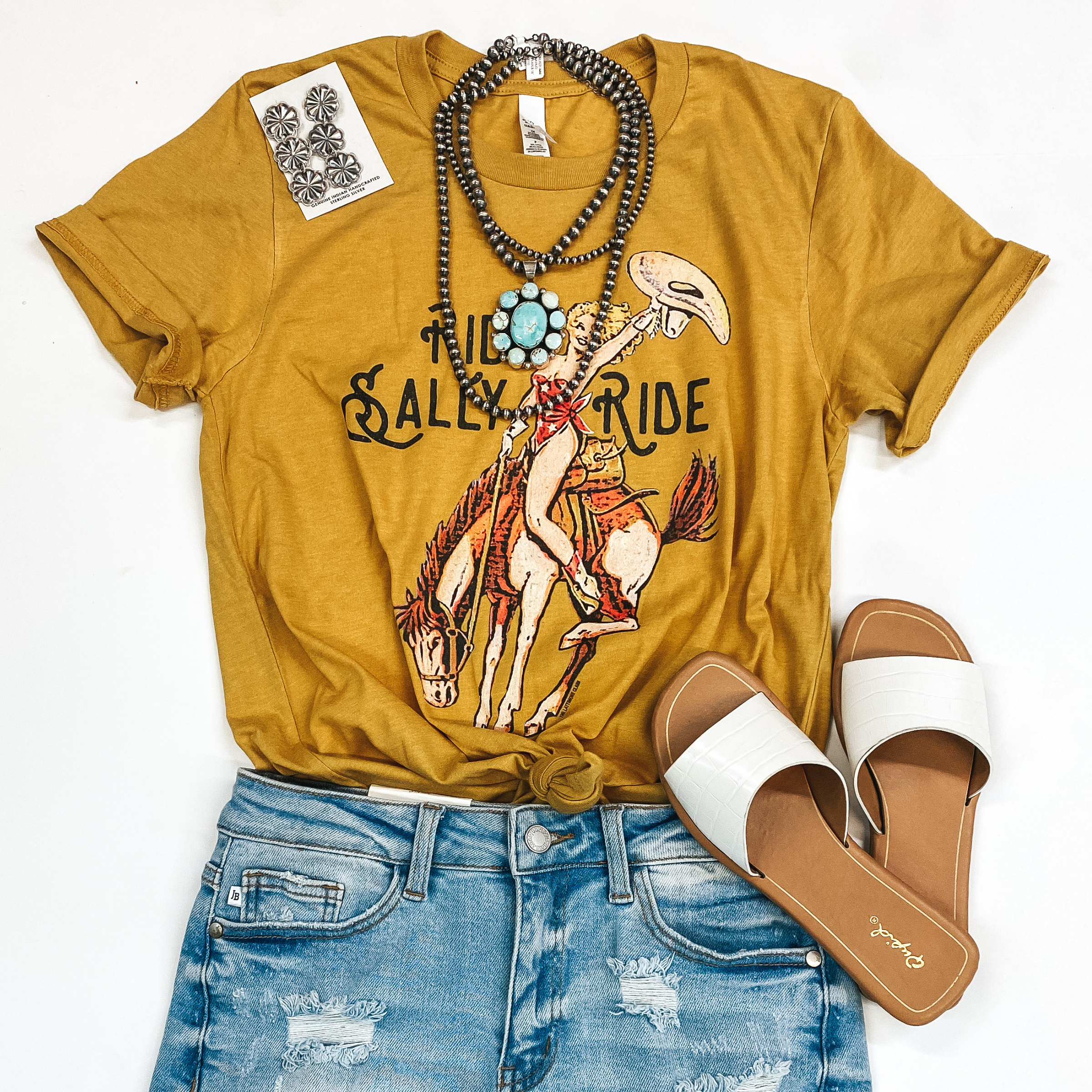 In the middle of the picture is a bucking horse with a woman on top holding a cowboy hat, in mustard yellow. This tee is paired with navajo jewelry, white sandals, and blue jean shorts. Background is solid white. 
