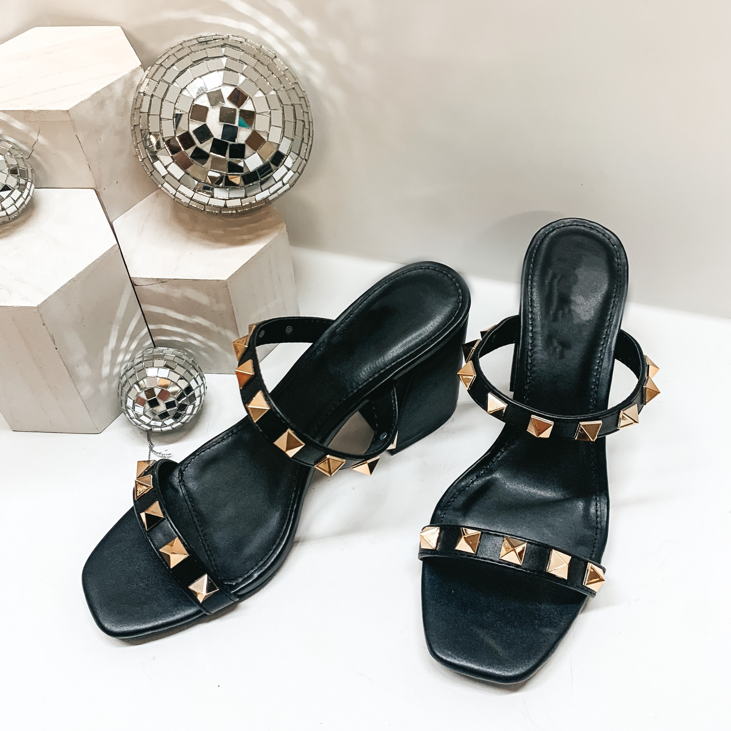 A pair of black strappy heels that have a square toe and block heel. The straps also have gold studs. These heels are pictured on a white background with white blocks and disco balls in one corner. 