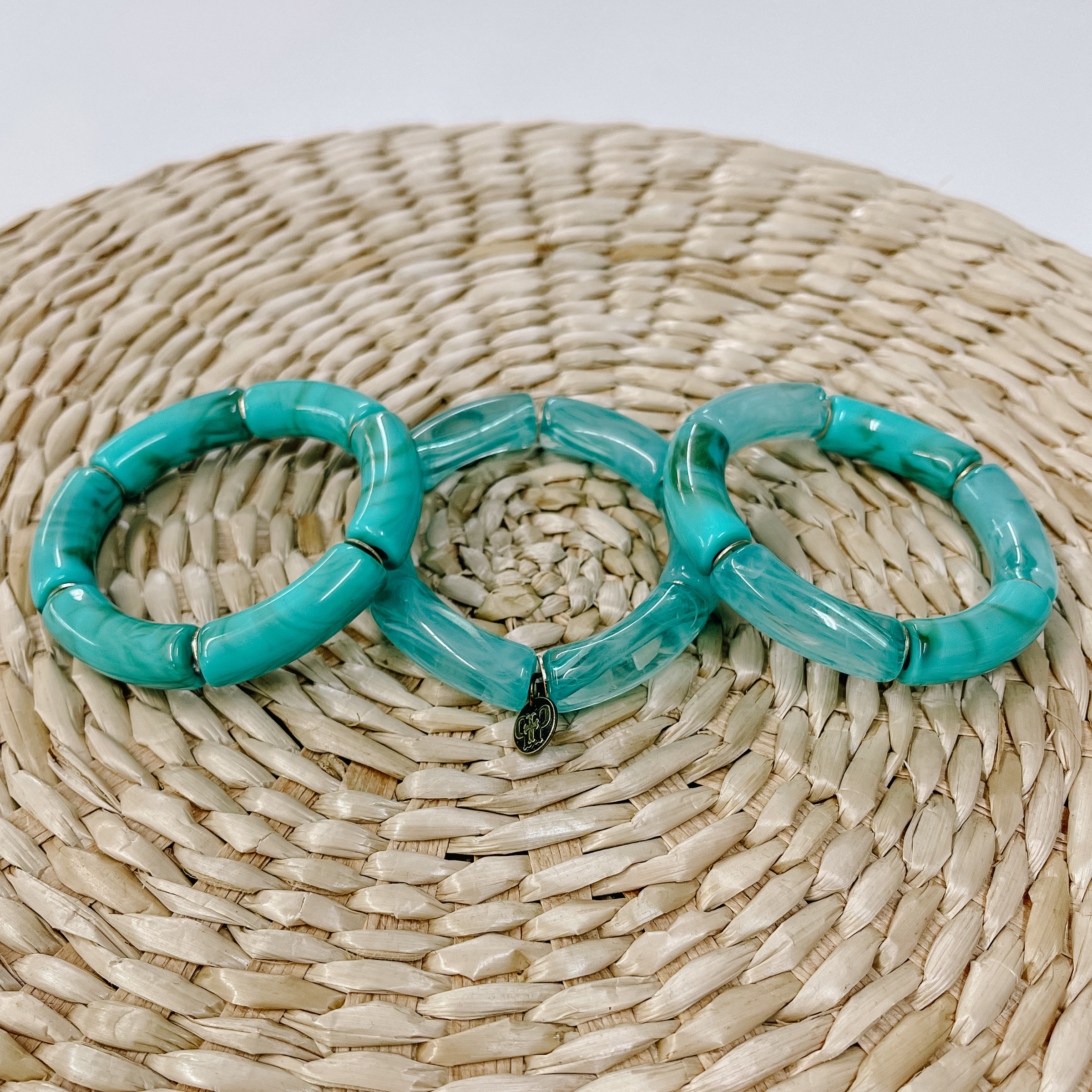 These are three acrylic tube bracelets in teal  with different acrylic patterns and gold spacers. The bracelet in the  left is a mix of solid teal and green marble. The middle one is clear teal marble tubes.  The right one is a mix of clear teal marble tubes and solid teal and green marble tubes. Taken on top of a bamboo stool and white background.