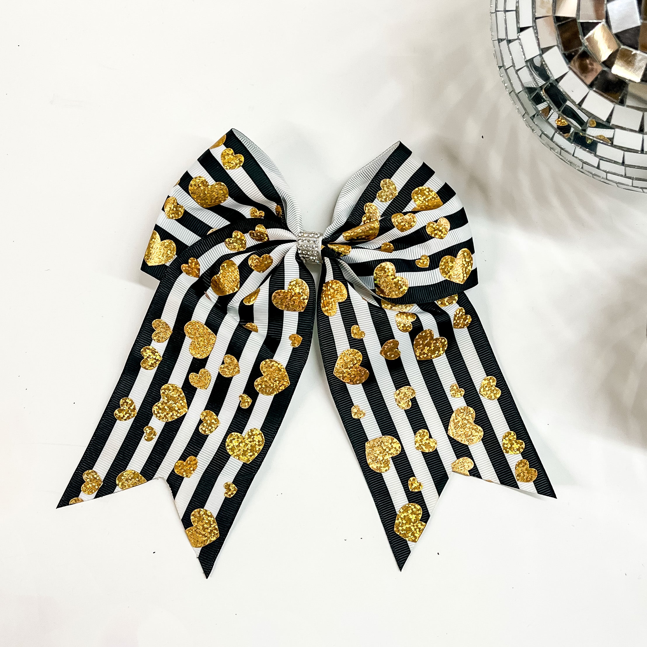 This is a black and white striped bow with gold hearts all over and the center of the bow has clear rhinestones. This bow is taken on a white background with a disco ball in the side as decor.