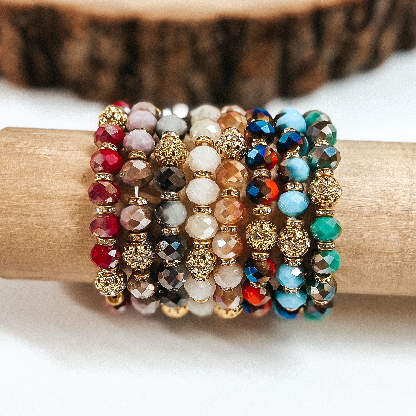 There are eight crystal beaded bracelets in different colors and textures taken on a bracelet wood holder. These bracelets are takekn on a white background with a slab of wood in the back as decor.