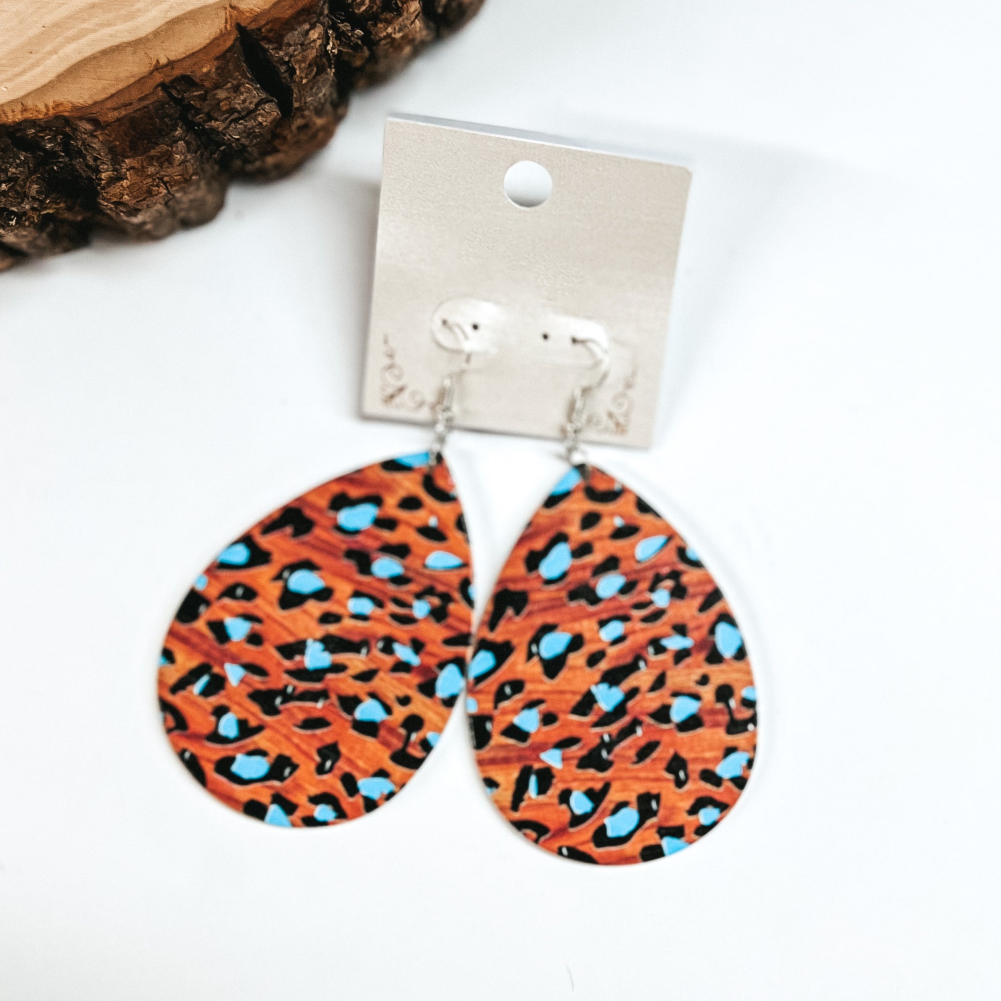 These are brown wooden teardrop earrings in leopard print and blue detailing. These earrings are placed on a white earring card holder, they are laying on a  white background with a slab of wood in the back as decor.