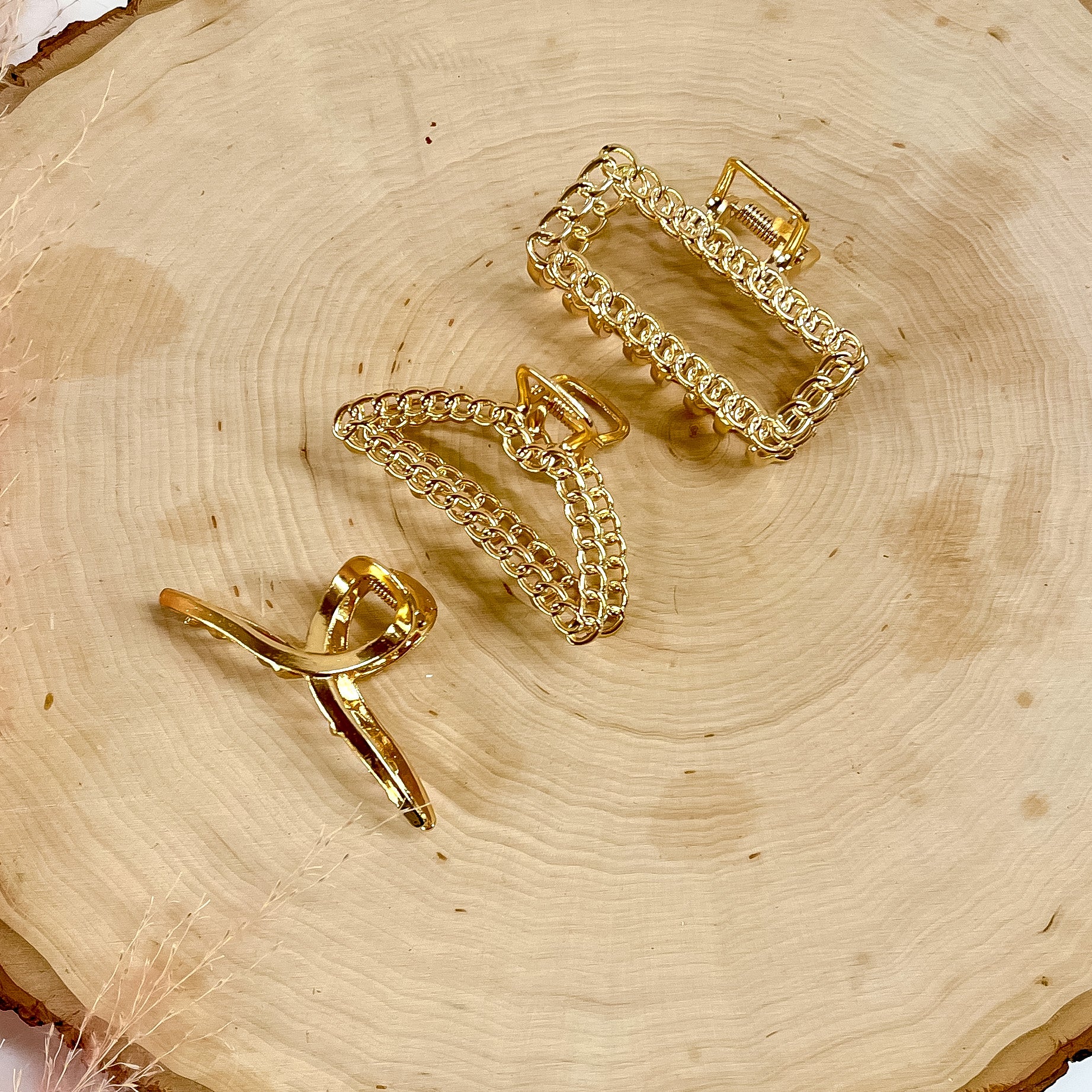 There are three gold small clips layingn on a slab of wood, all three clips have different textures and shapes. From top to bottom; rectangle chain , dome chain texturesd, and smooth loop.