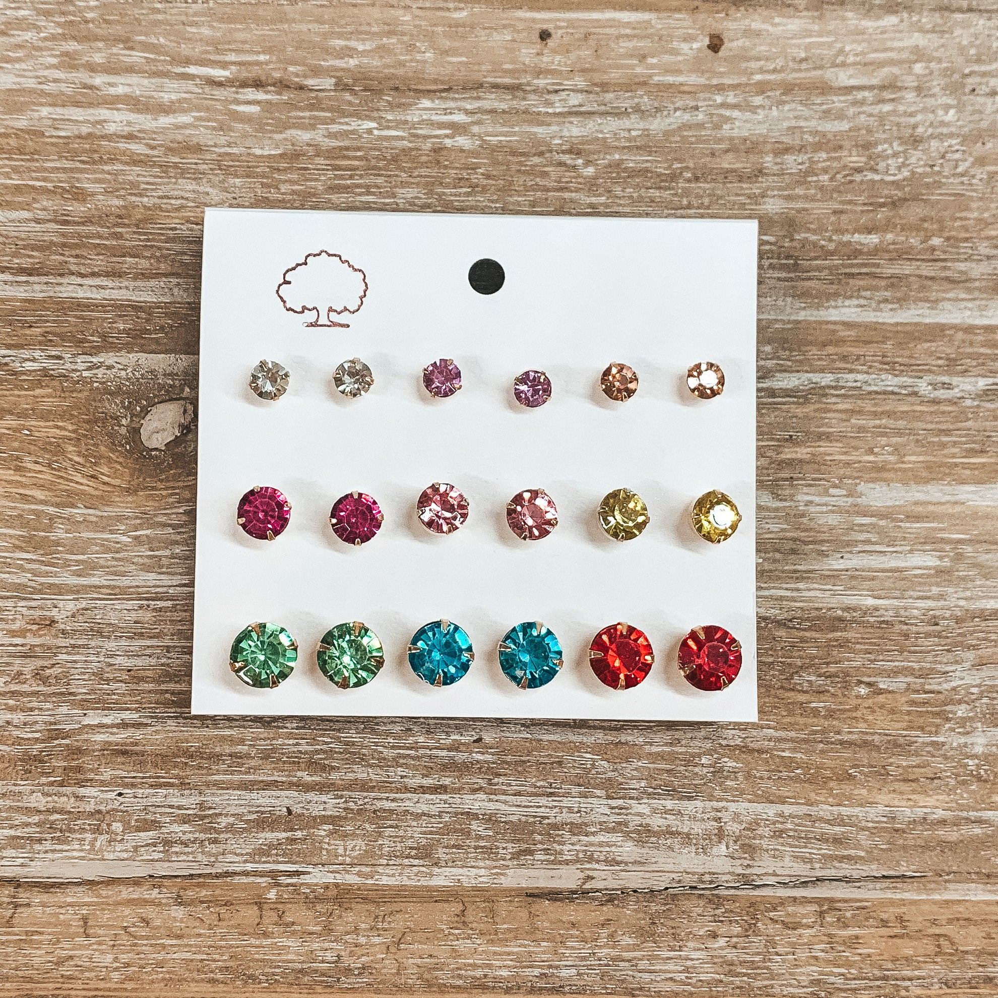 This is a pack of nine multicolored crystal stud earrings in clear, pink, yellow, green, blue, and red. These earrings are on a white earring card holder and taken on a wooden slate.