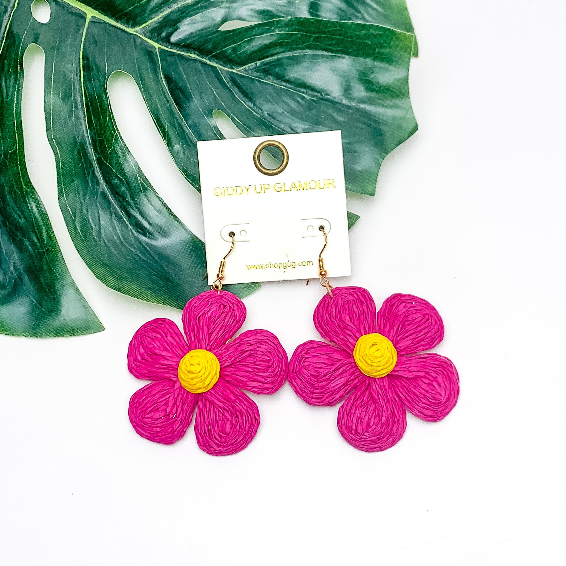 Darling Daisy Raffia Wrapped Flower Earrings in Hot Pink. Pictured on a white background with the earrings laying on a large leaf.