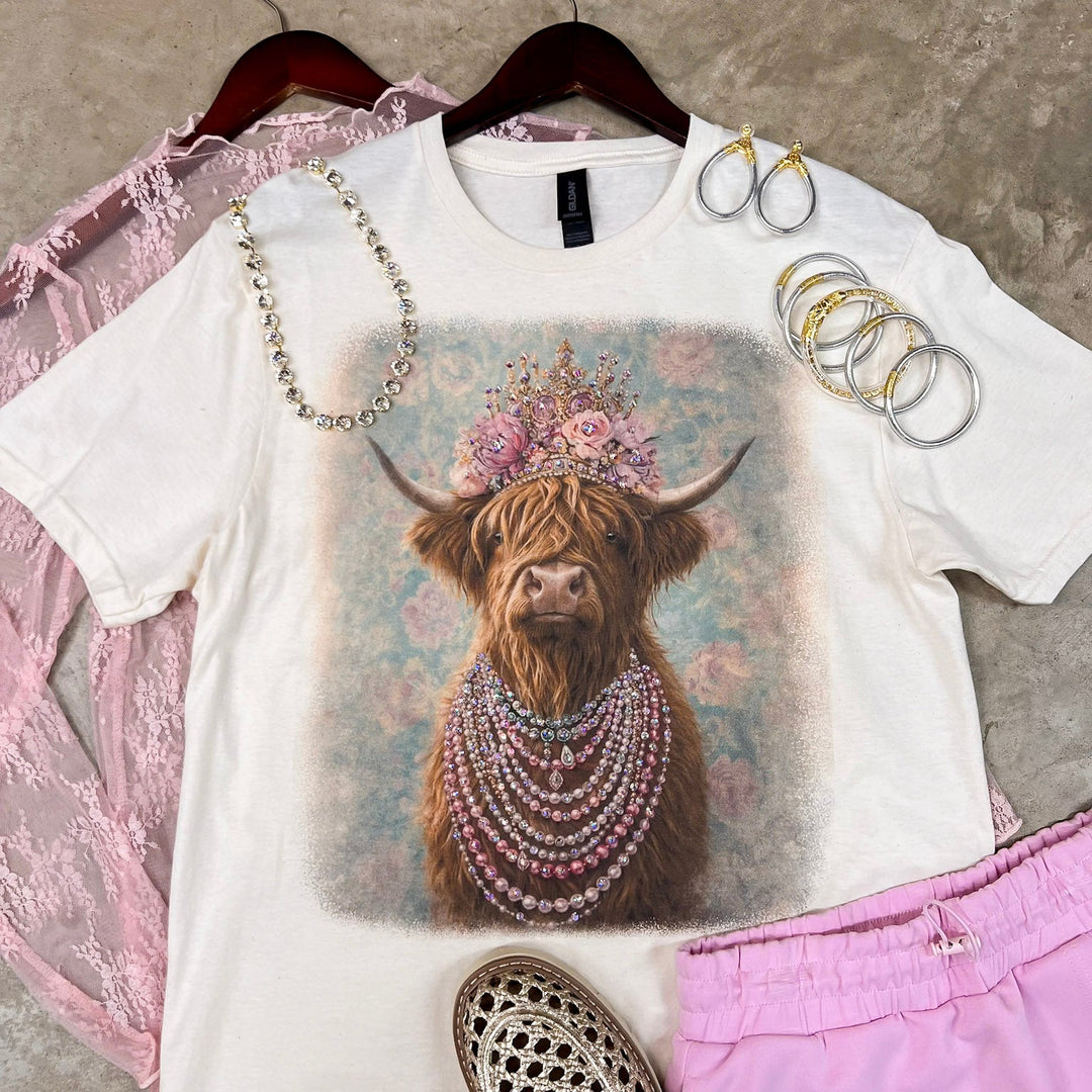 A styled flat-lay of a white graphic T-shirt on a hanger, featuring a highland cow illustration with a floral crown and layered pearl necklaces. The shirt is styled with pink lace fabric, silver hoop earrings, gold rings, metallic loafers, and pink shorts on a textured concrete floor.