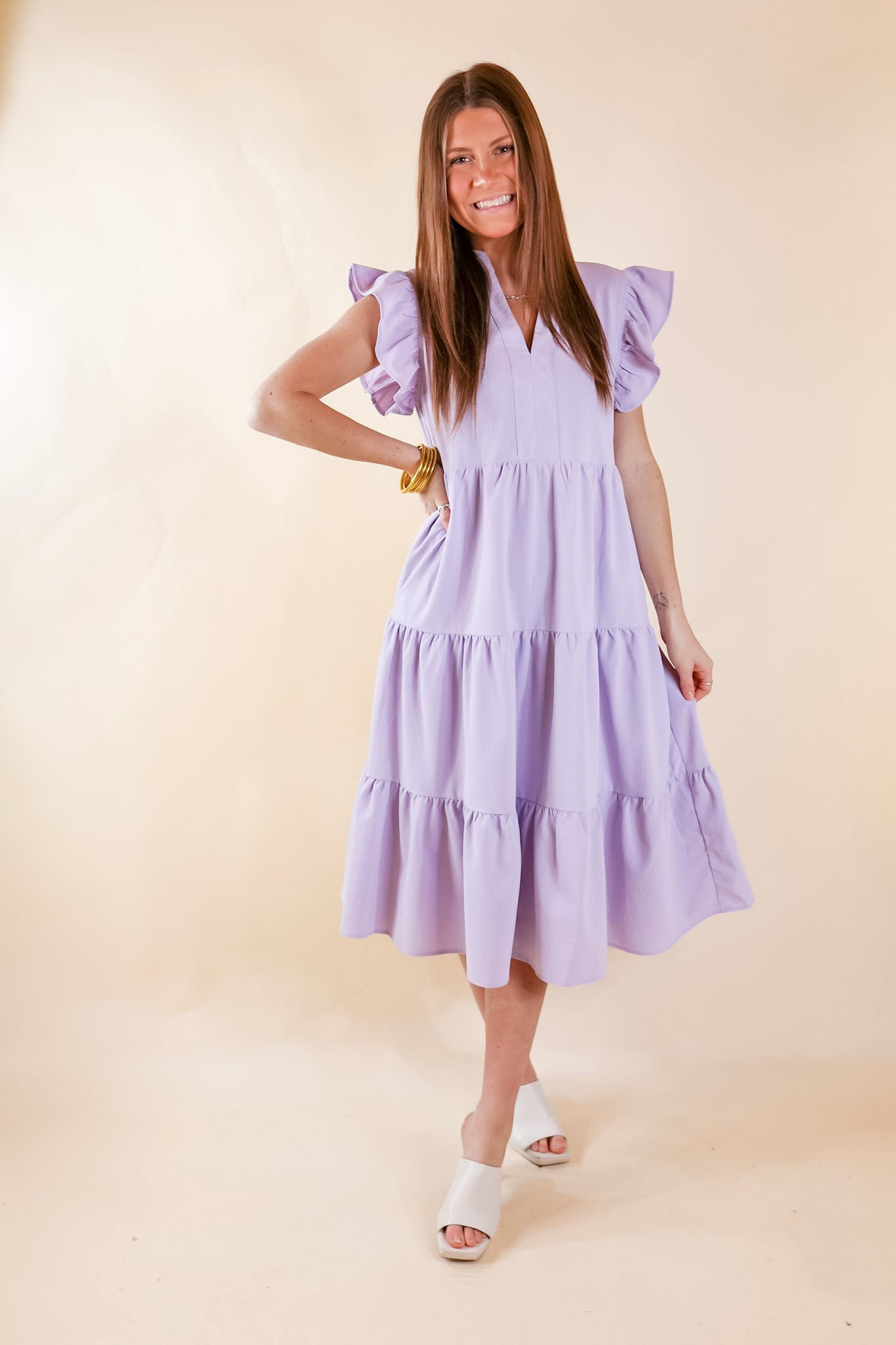A relaxed fit midi dress in lavender. The dress has a V neckline, short ruffled sleeves, pockets, and a tiered skirt. Item is pictured on a pale pink background