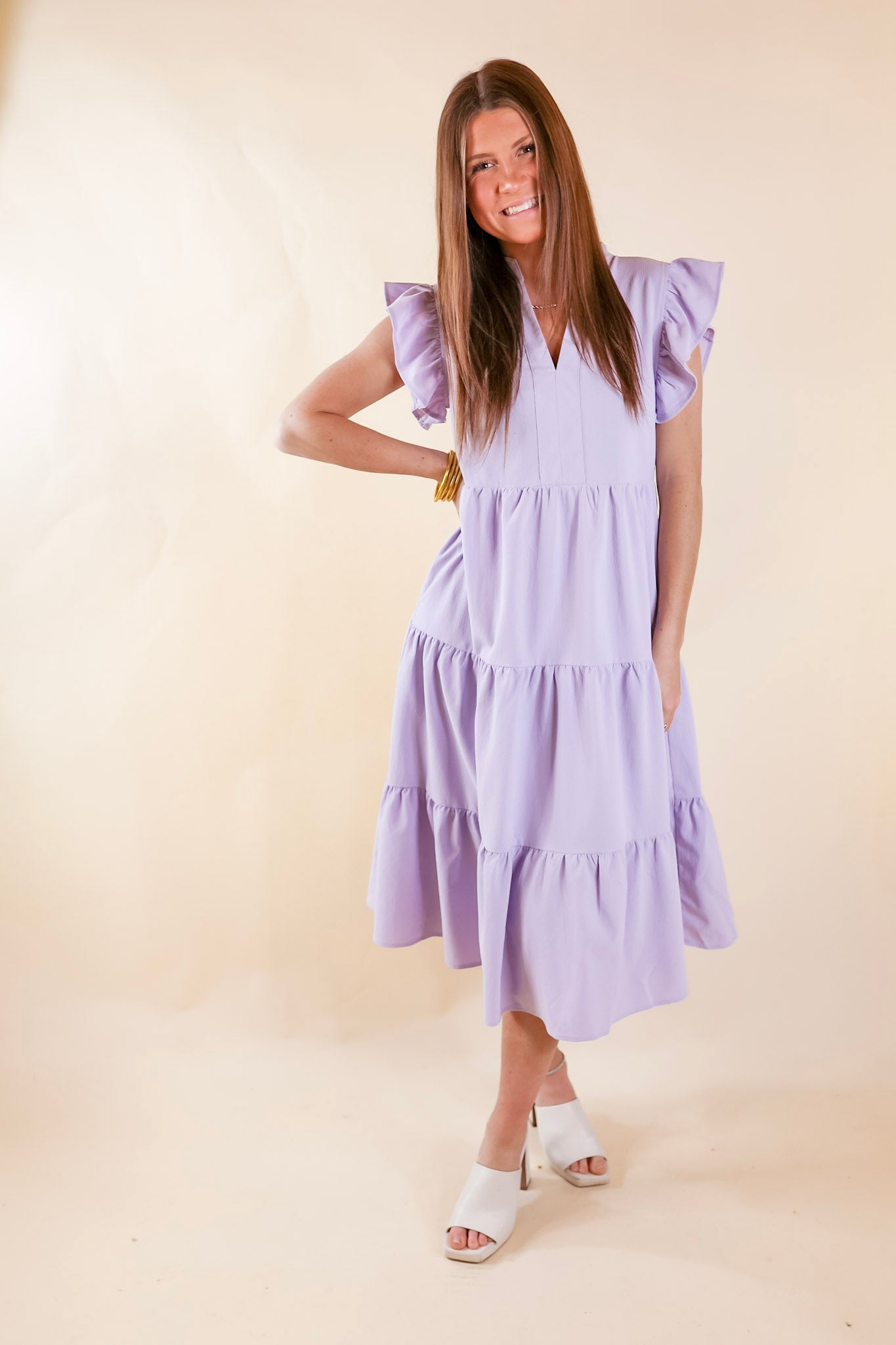 A relaxed fit midi dress in lavender. The dress has a V neckline, short ruffled sleeves, pockets, and a tiered skirt. Item is pictured on a pale pink background