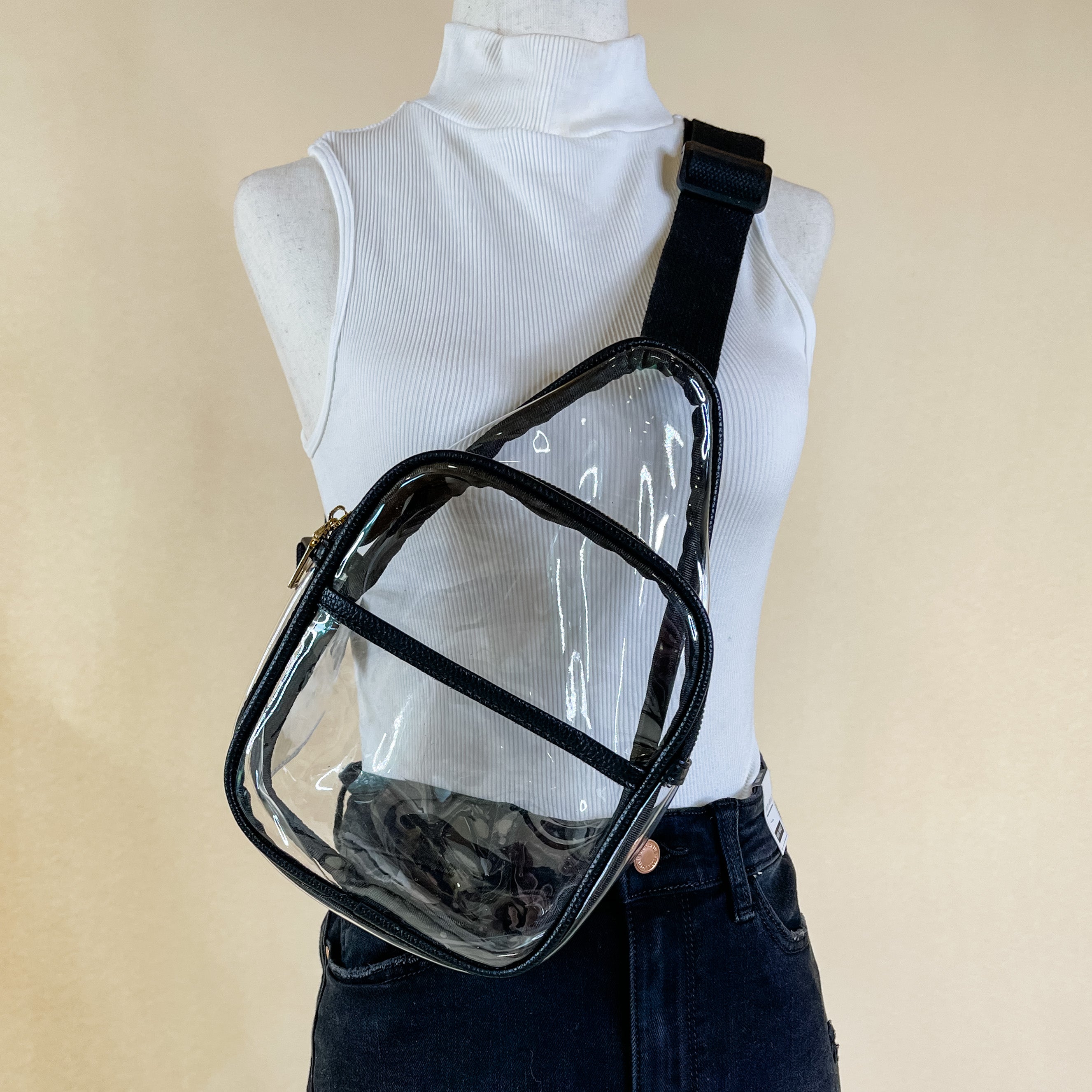Pictured is a clear sling backpack with a black outline. This bag also includes a black strap and black and gold accents. This bag is on a mannequin that has black shorts and a white high neck tank pictured in front of an ivory background.  