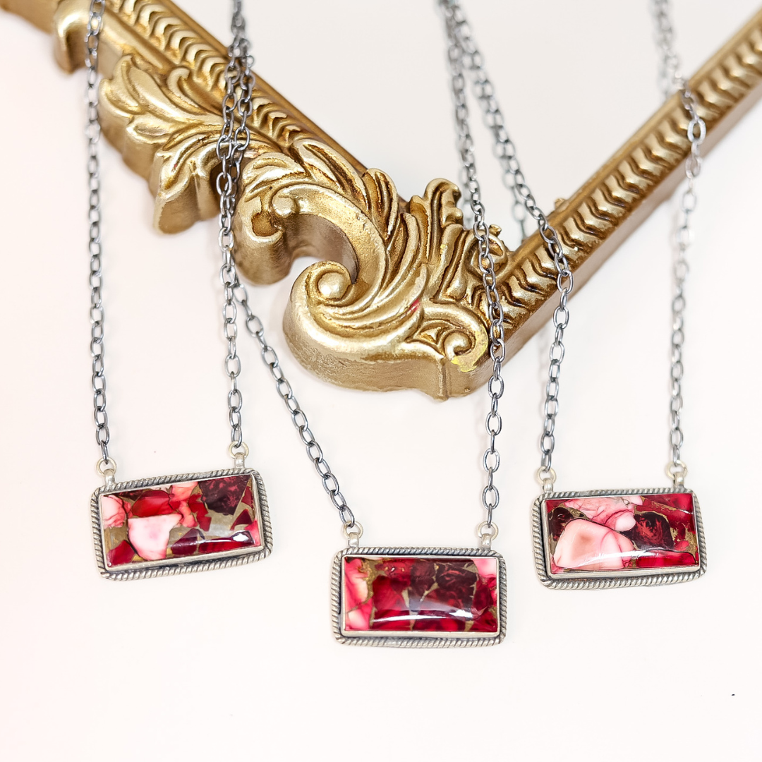 Close-up of handcrafted Rose Dahlia bar necklaces featuring pink and red stones set in sterling silver on 20-inch oxidized chains, displayed on ornate gold frame.