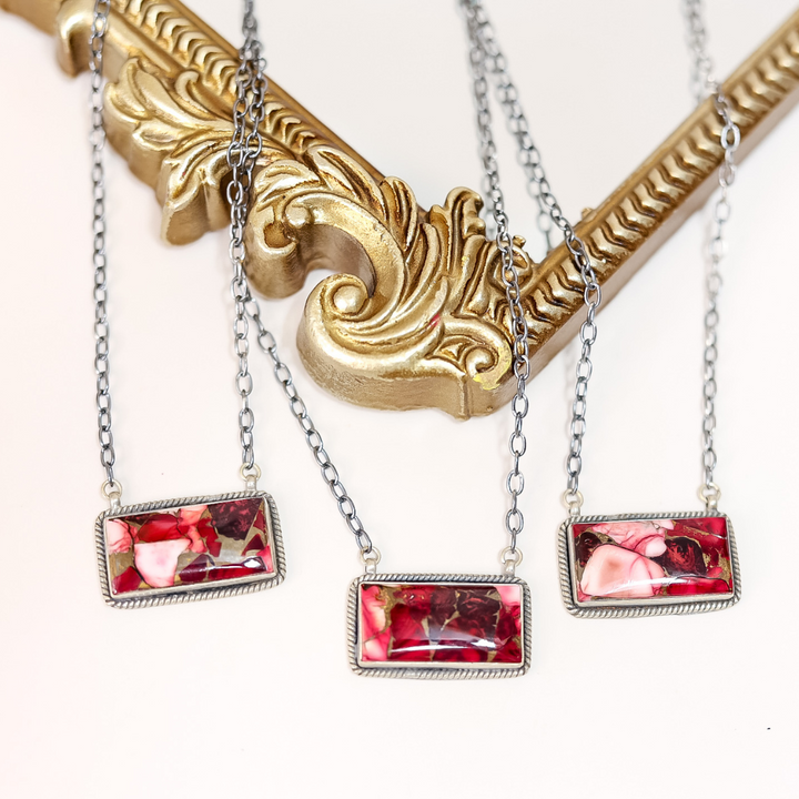 Close-up of handcrafted Rose Dahlia bar necklaces featuring pink and red stones set in sterling silver on 20-inch oxidized chains, displayed on ornate gold frame.