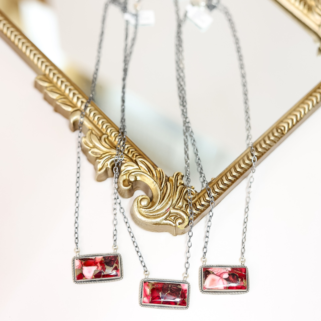 Close-up of handcrafted Rose Dahlia bar necklaces featuring pink and red stones set in sterling silver on 20-inch oxidized chains, displayed on ornate gold frame.