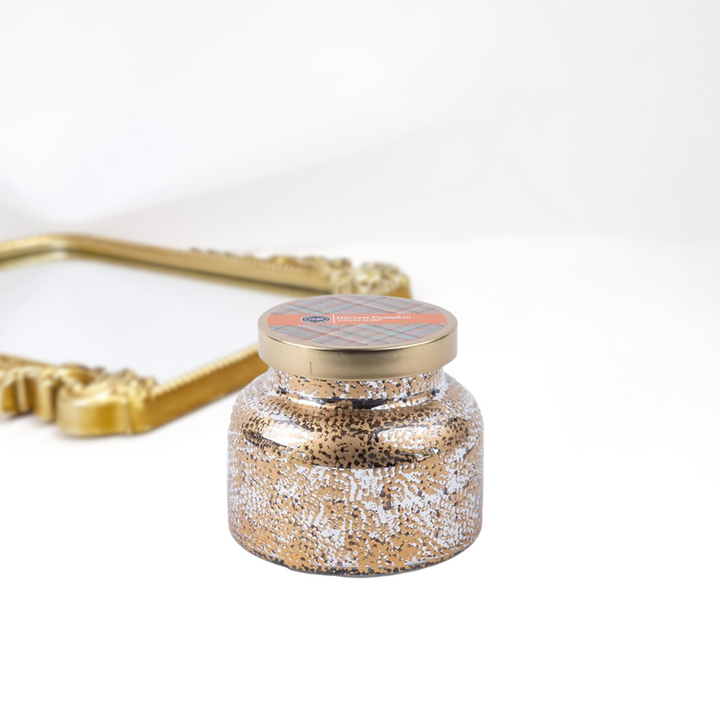 A solid white background with a gold mirror. A holiday inspired candle jar that is textured and has a gold lid.