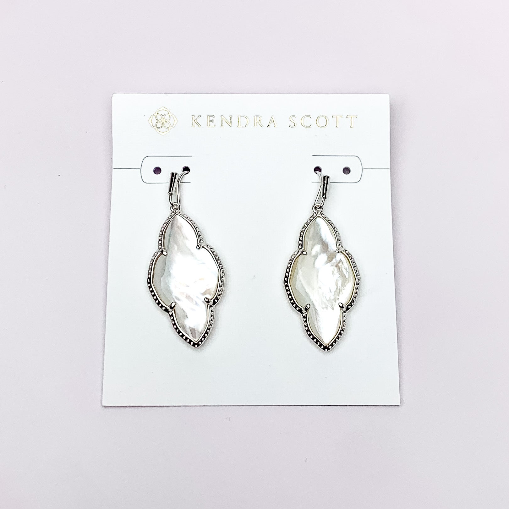 Silver drop earrings in a quatrefoil shape with an ivory mother of pearl stone. These earrings are pictured on a white earring holder on a white background. 