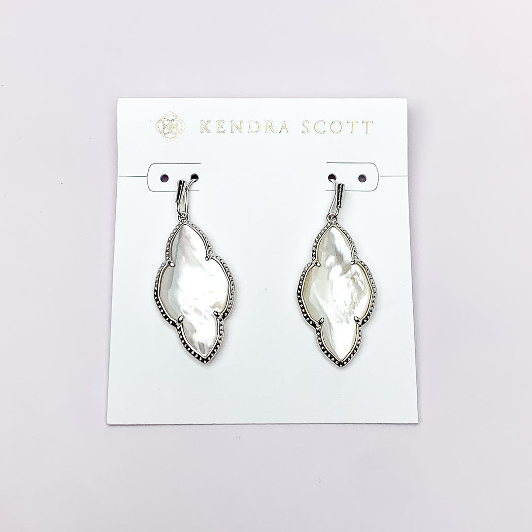 Silver drop earrings in a quatrefoil shape with an ivory mother of pearl stone. These earrings are pictured on a white earring holder on a white background. 