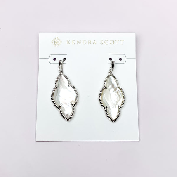 Silver drop earrings in a quatrefoil shape with an ivory mother of pearl stone. These earrings are pictured on a white earring holder on a white background. 
