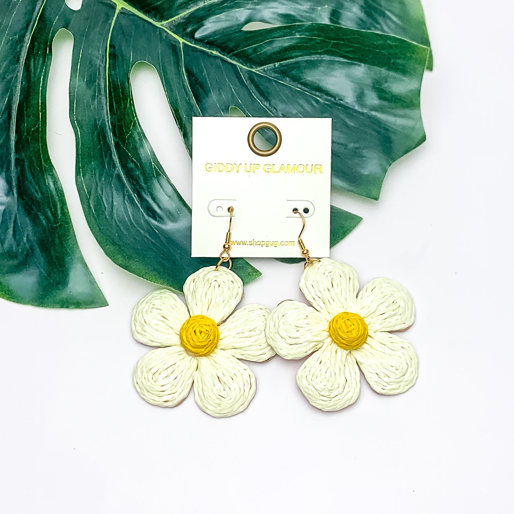 Darling Daisy Raffia Wrapped Flower Earrings in Ivory. Pictured on a white background with the earrings laying on a large leaf.