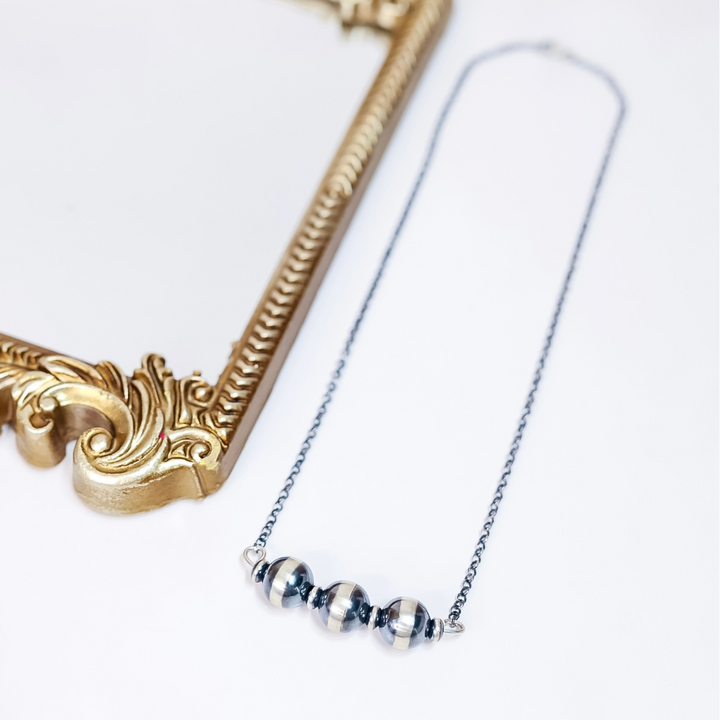 Close-up of Navajo Pearl Bar Necklace with 16mm sterling silver beads and saucer spacers on a 20-inch oxidized chain, styled flat with gold frame accent.