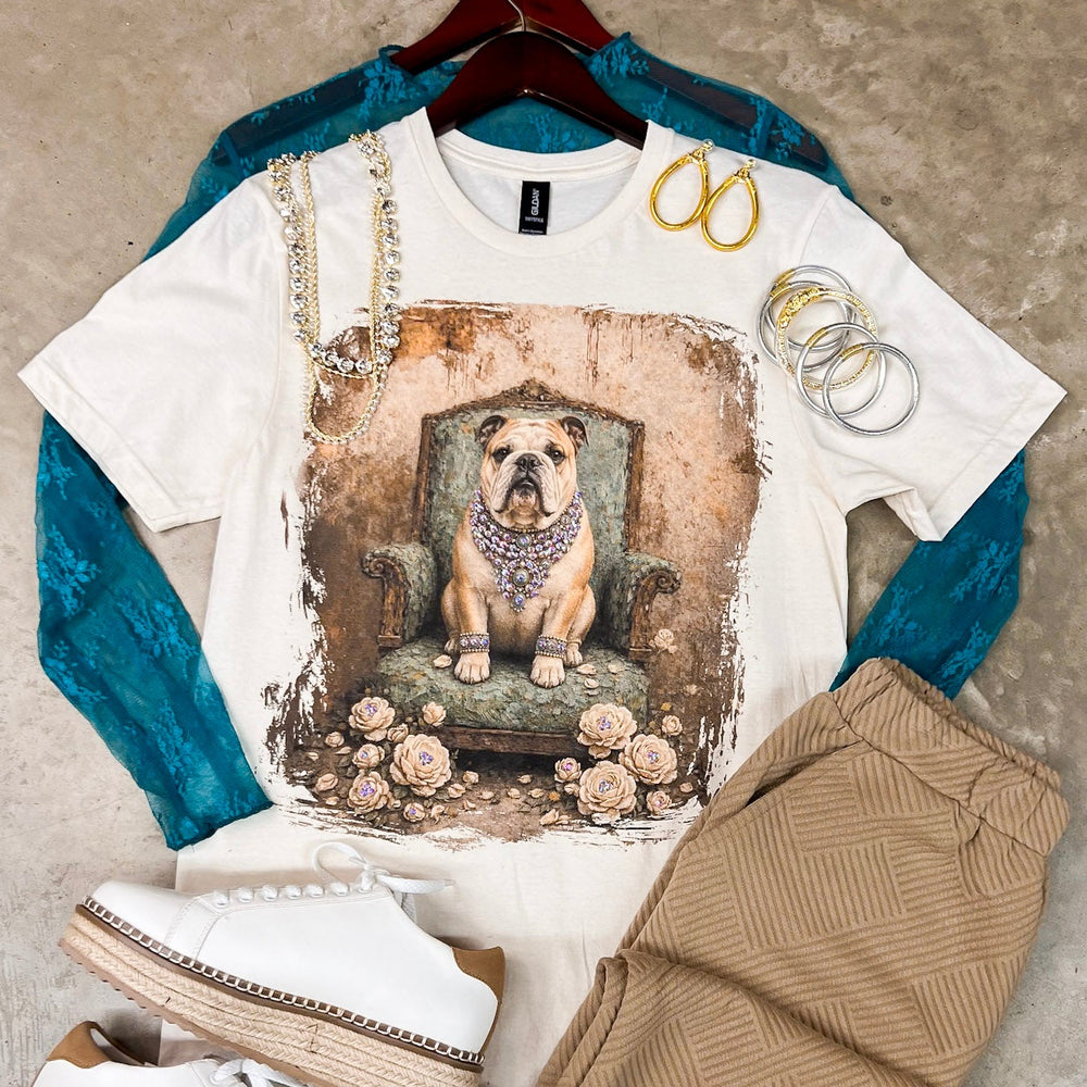 A styled flat-lay outfit featuring a white graphic t-shirt with a bulldog print, layered over a teal long-sleeve top, paired with tan herringbone shorts, white platform espadrille sneakers, and gold jewelry, all arranged on a textured concrete floor.