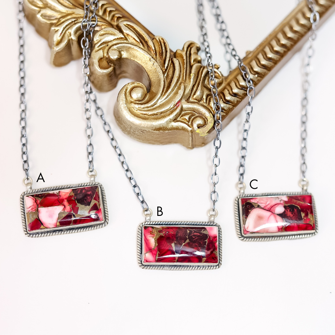 Close-up of handcrafted Rose Dahlia bar necklaces featuring pink and red stones set in sterling silver on 20-inch oxidized chains, displayed on ornate gold frame.