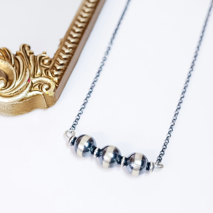 Close-up of Navajo Pearl Bar Necklace with 16mm sterling silver beads and saucer spacers on a 20-inch oxidized chain, styled flat with gold frame accent.