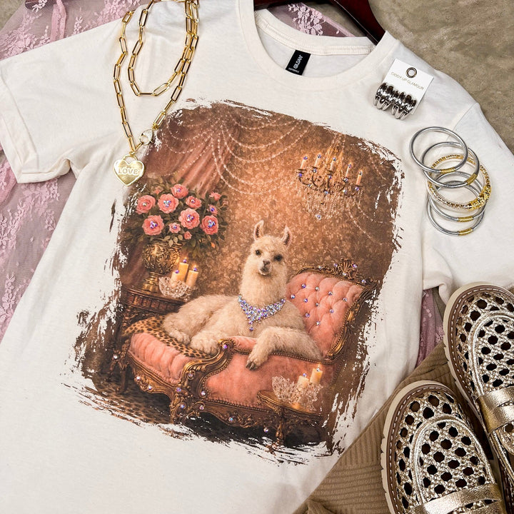 A white graphic T-shirt laid flat on a hanger, featuring a vintage-style print of a small dog lounging on a pink chaise lounge with flowers and ornate details. The shirt is styled with gold necklaces, silver rings, a pink lace garment, and metallic loafers on a textured beige floor.