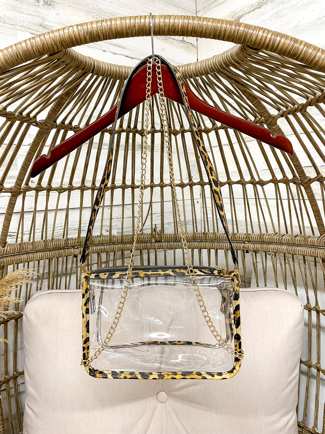 Clear handbag with leopard print trim hanging on a red hanger against a wicker chair background.